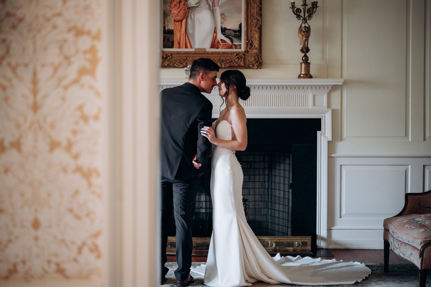 Bride and groom embracing tenderly in elegant white fireplace room during wedding day portraits.