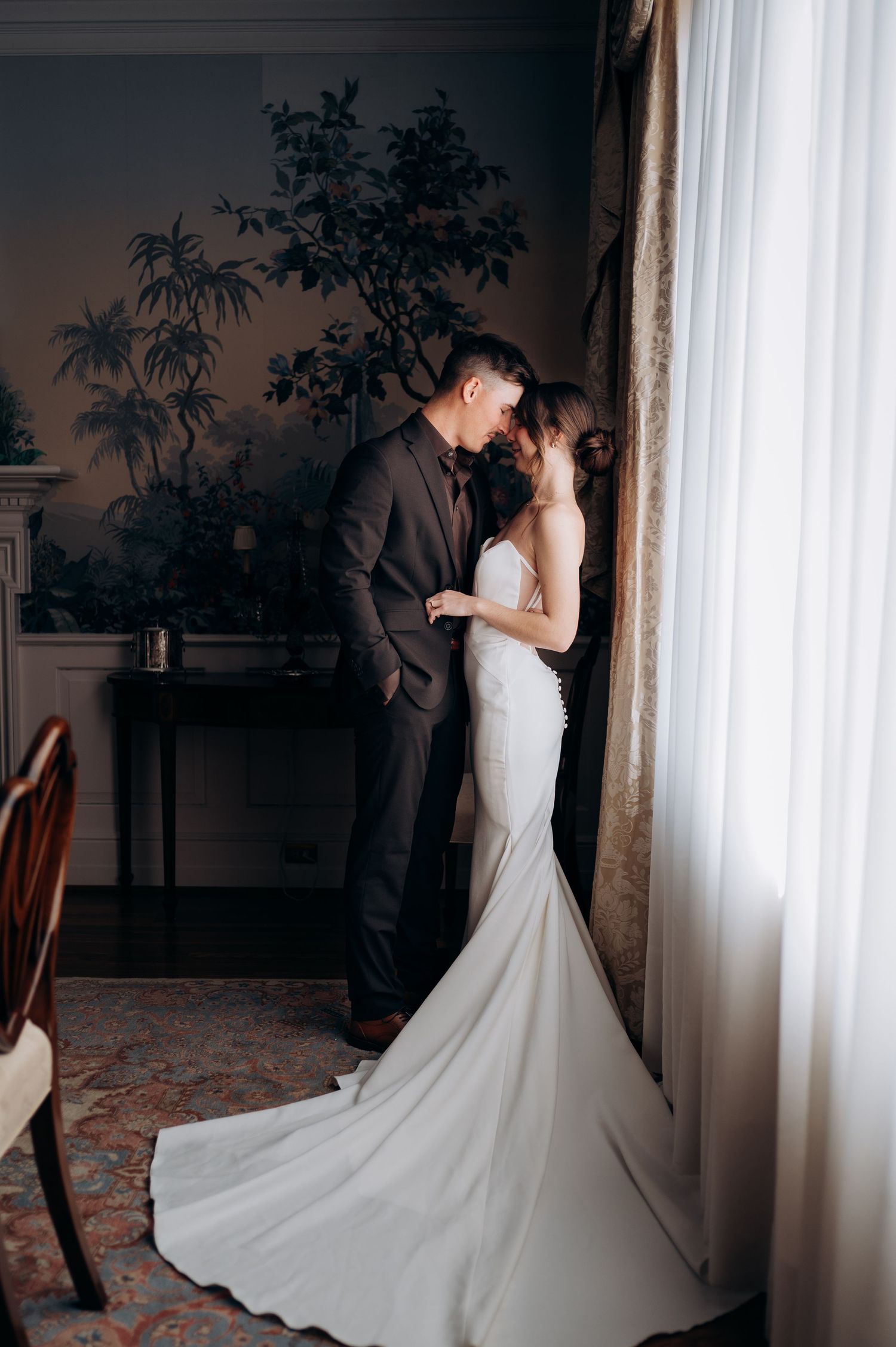 Elegant bride in fitted white mermaid gown with train stands with groom in brown suit by sheer curtained window.