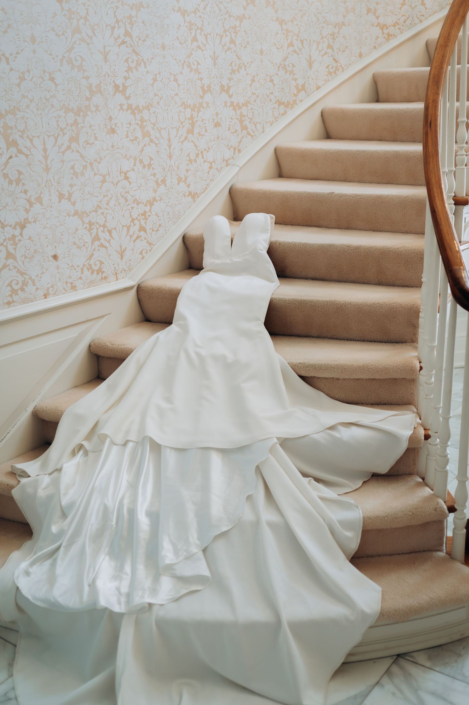 Elegant white wedding gown with layered skirt and long train draped over carpeted staircase in classic interior.