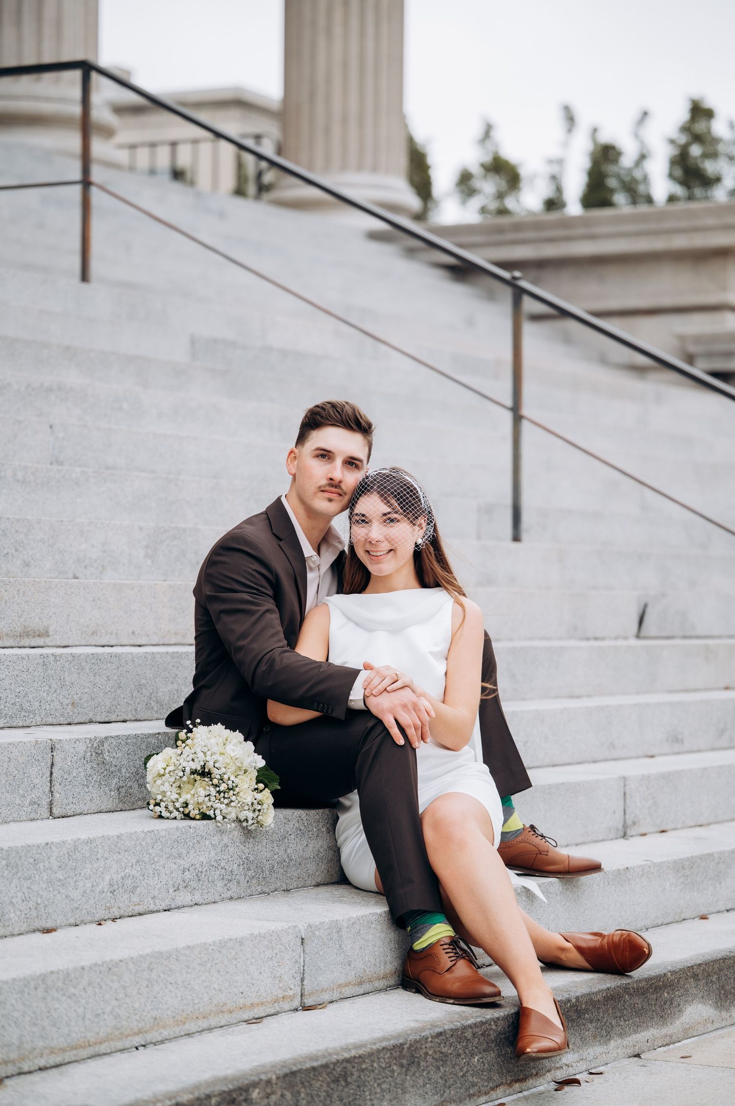 Newlyweds sitting together on marble steps, bride in white dress with birdcage veil, bouquet beside them.