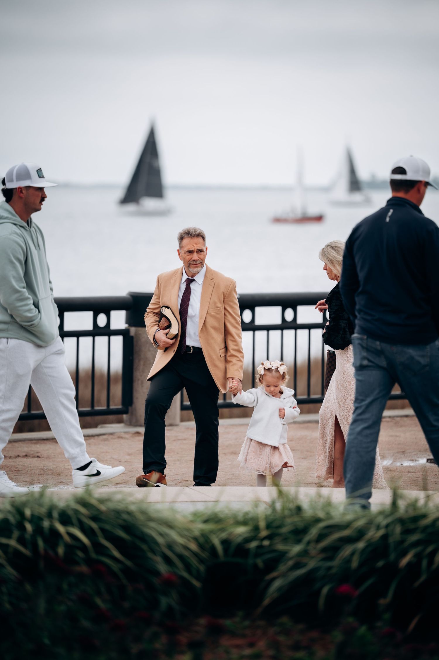 Man in tan blazer holds hands with toddler girl near waterfront with sailboats in background.