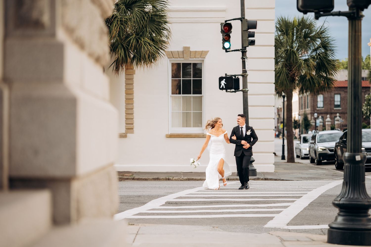 Bride and groom cross a sunny street in Charleston, SC, bride holding a white bouquet.