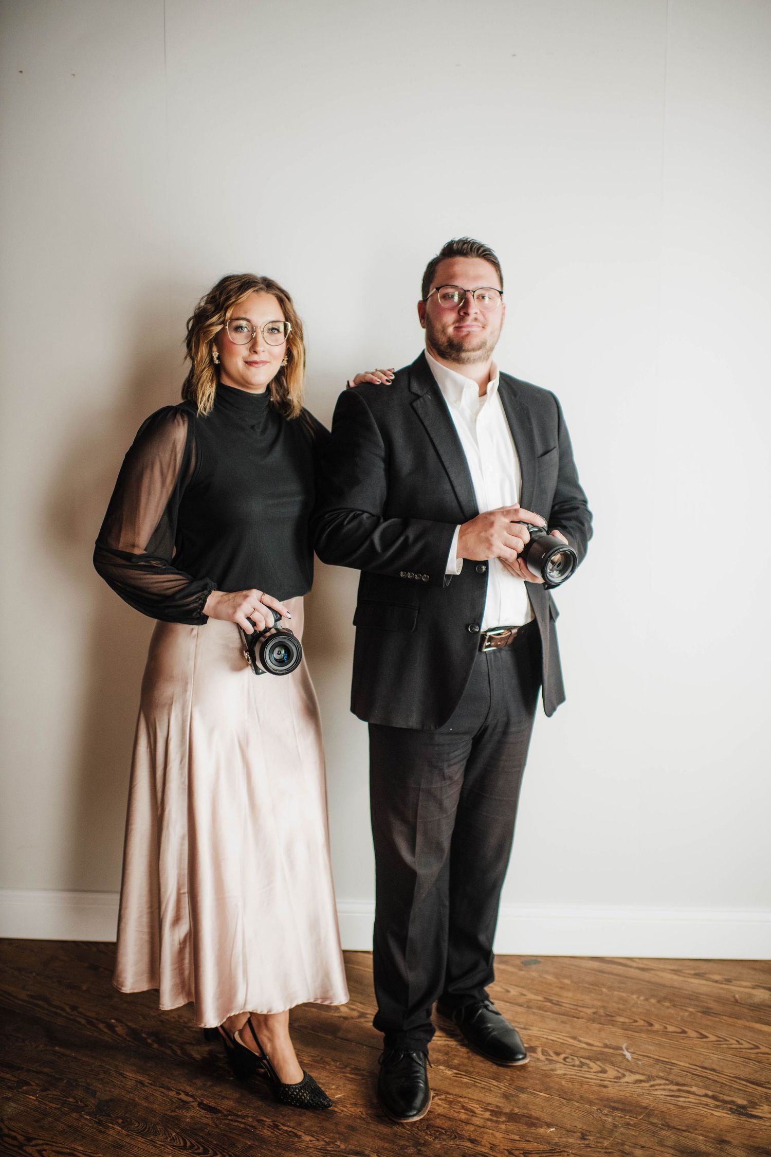 Two professional photographers in formal attire holding cameras pose together against a white wall.