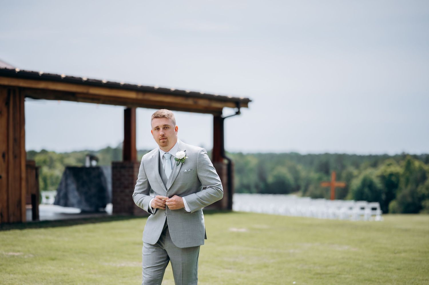 Groom in a light gray suit standing outdoors at a wedding venue with white chairs and a wooden structure in the background.