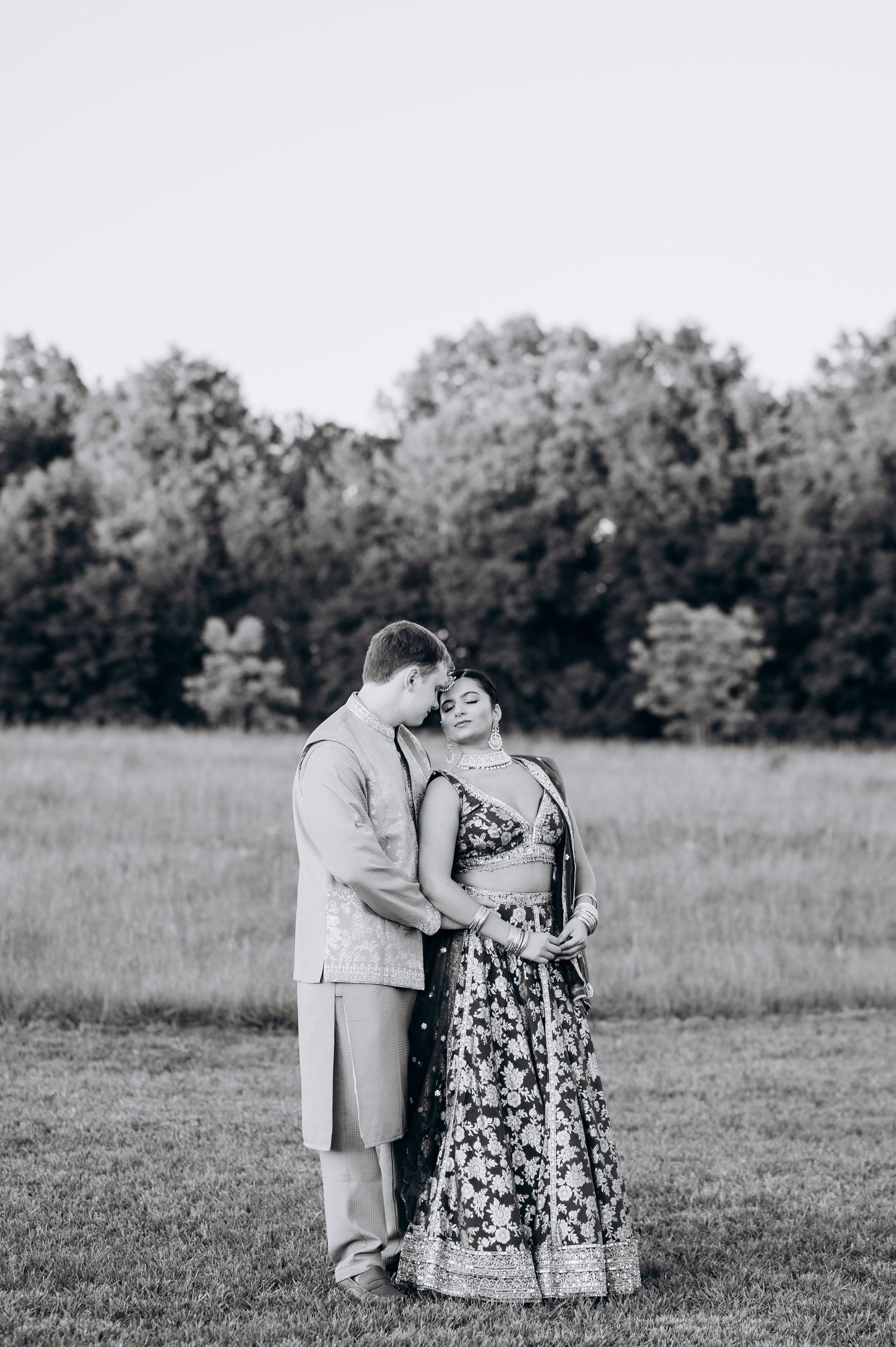 Black and white photo of a couple in traditional Indian wedding attire embracing in a lush green field.