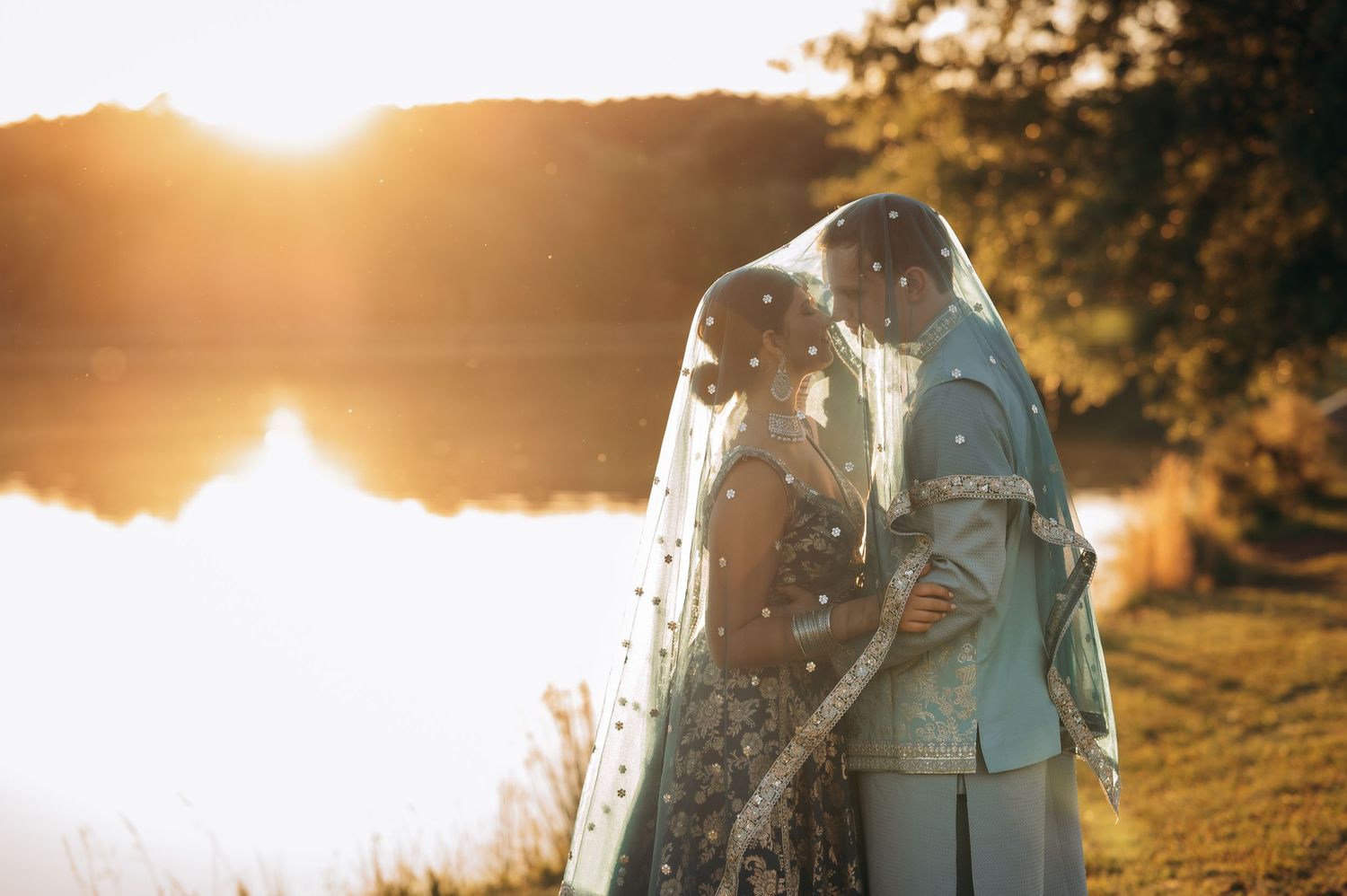 South Asian couple sharing romantic sunset kiss by lake, bride in embroidered veil, groom in blue sherwani.