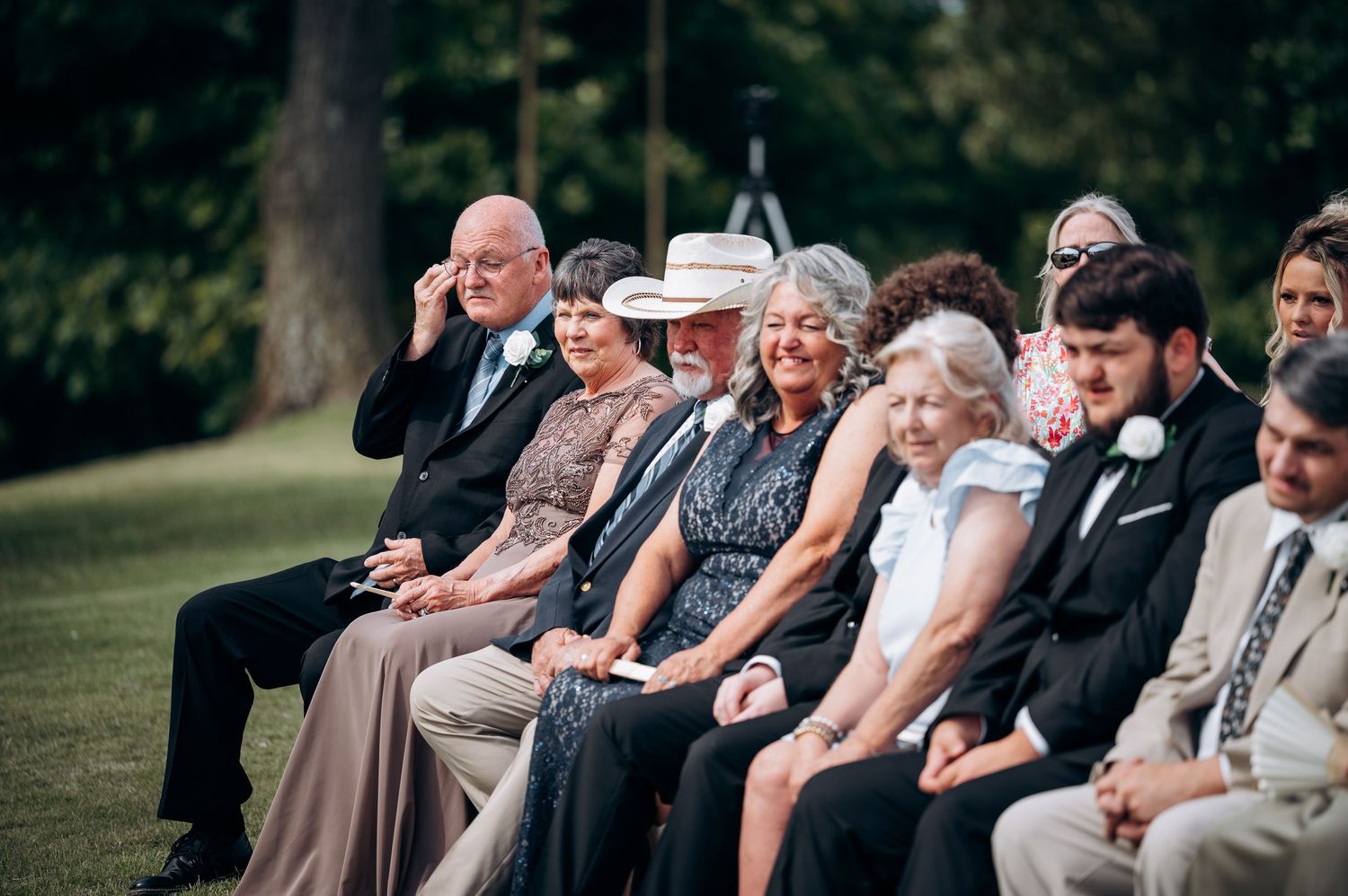 Wedding guests seated outdoors smile and watch the ceremony on a sunny day surrounded by trees.