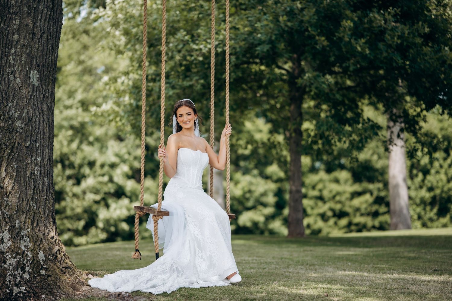 Bride in white wedding gown sitting on a rope swing under a large tree in a lush green garden.