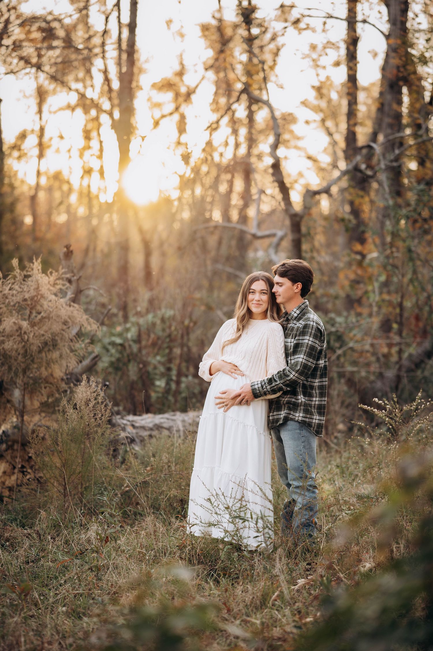 Couple embraces in sunlit forest during maternity photoshoot.