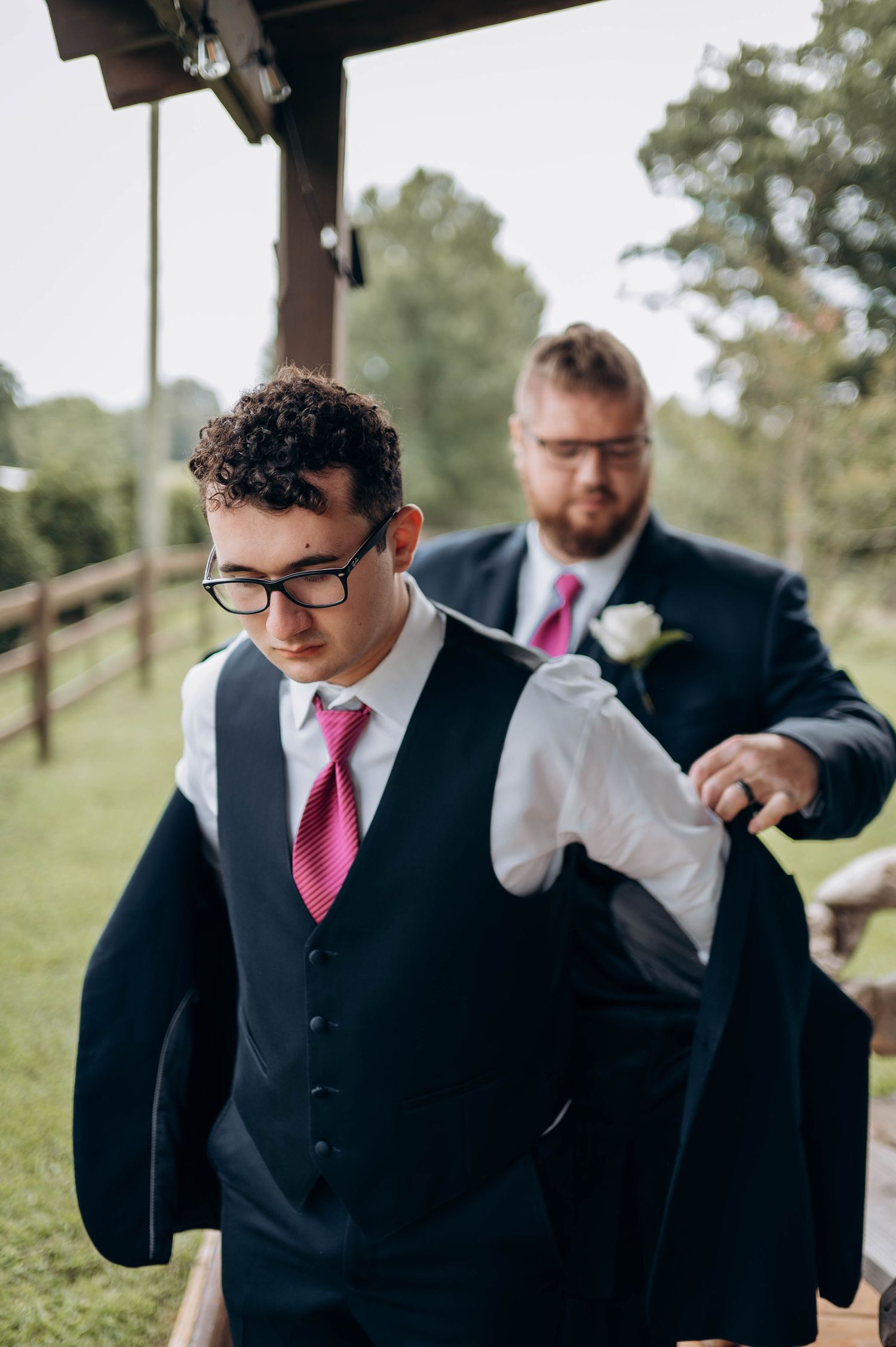 Groomsmen in black vests and pink ties preparing for wedding ceremony outdoors.
