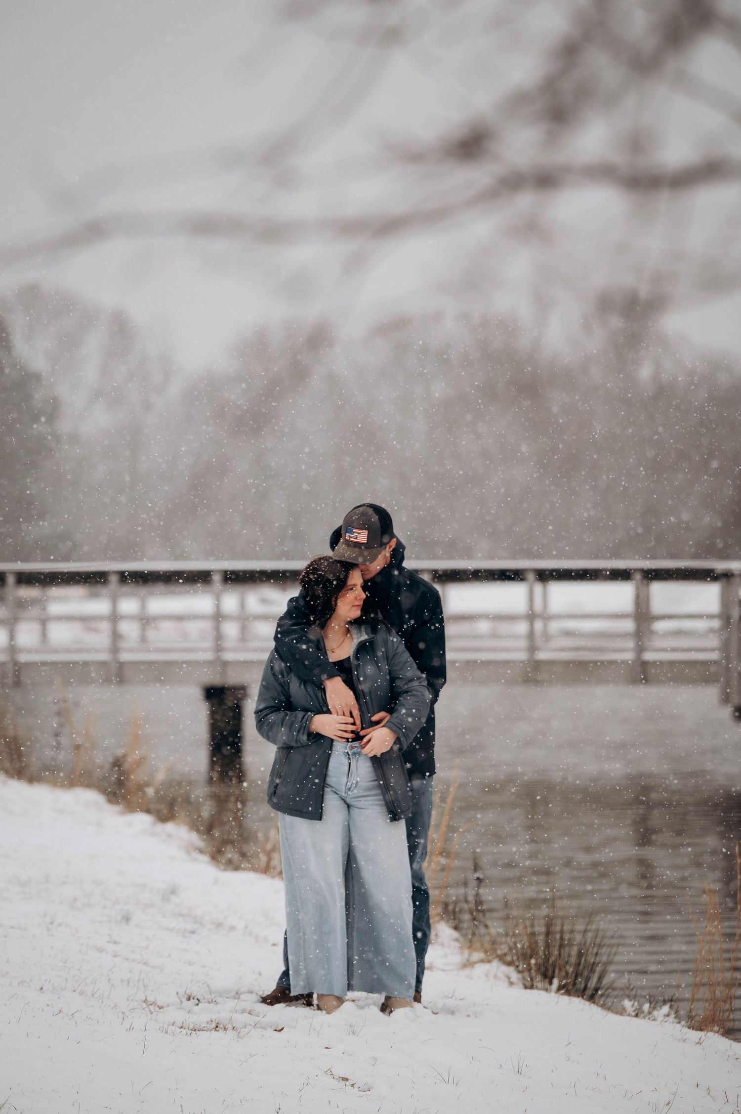 A romantic winter scene shows a couple embracing in the snowy landscape with a wooden fence in the background.