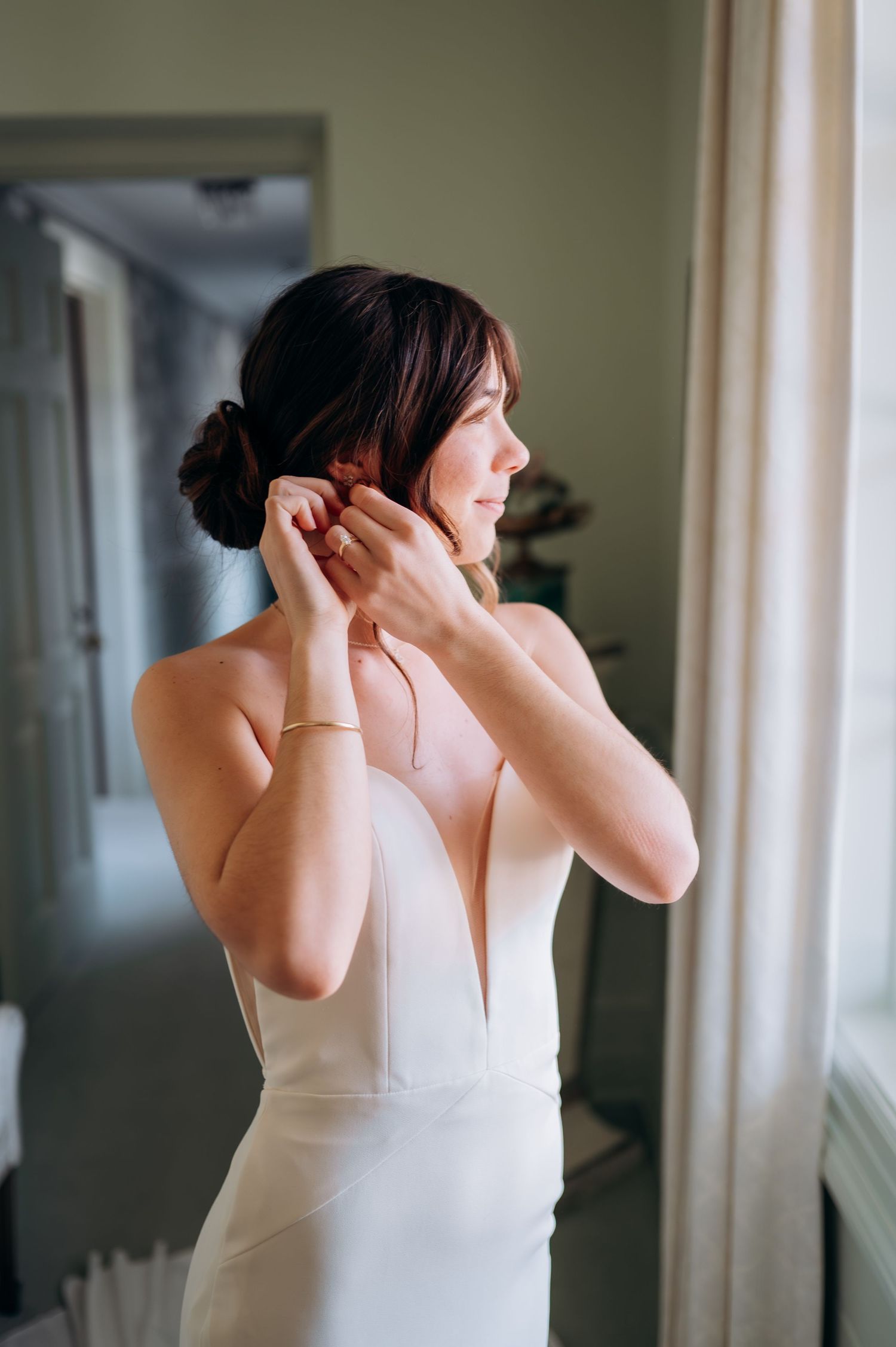 A woman in a white plunging neckline wedding dress puts on earrings while getting ready in a hallway.