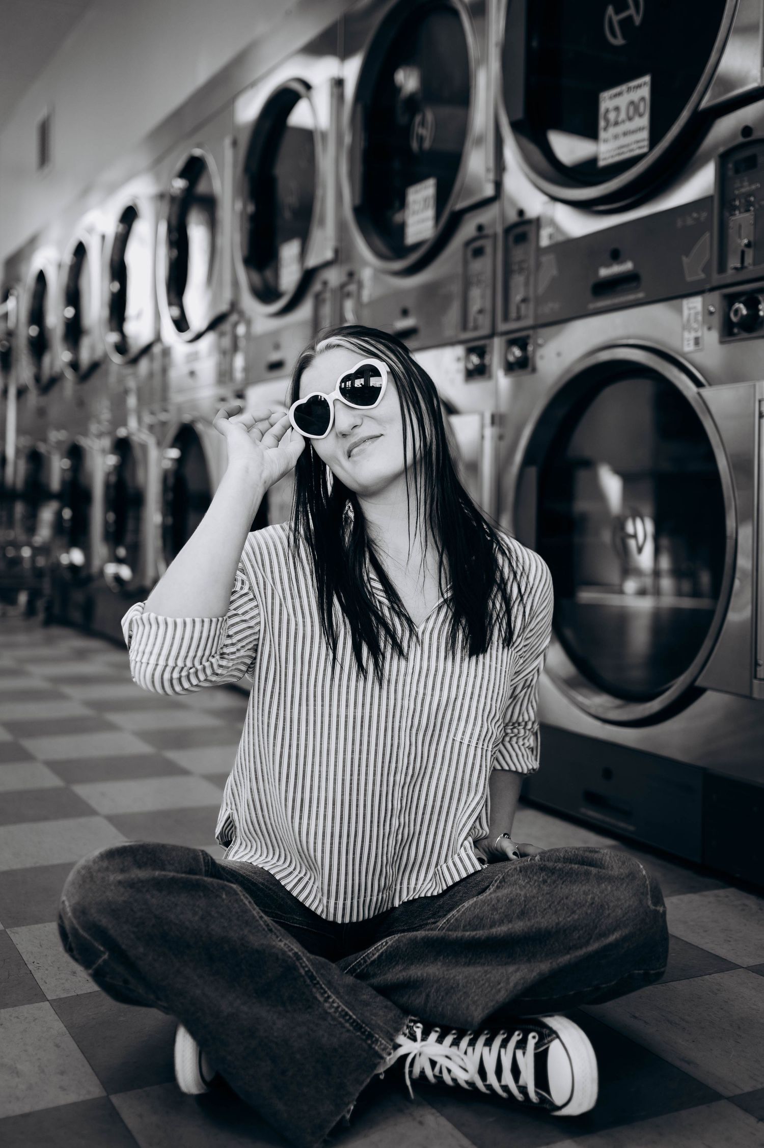Person in striped shirt and sunglasses sitting cross-legged in front of laundromat washing machines.