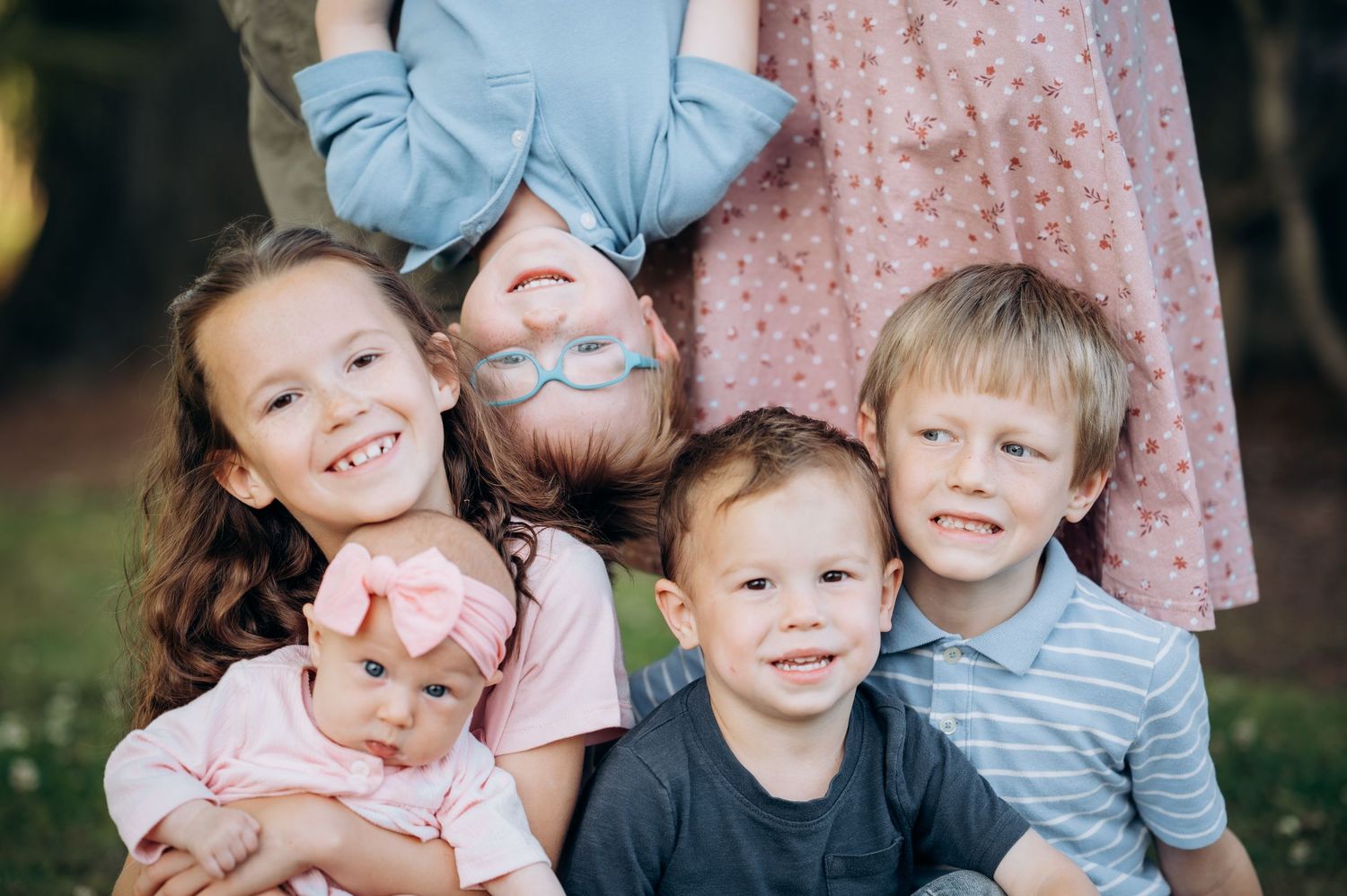 Five young children pose together outdoors, smiling for a family portrait in a lush green park setting.