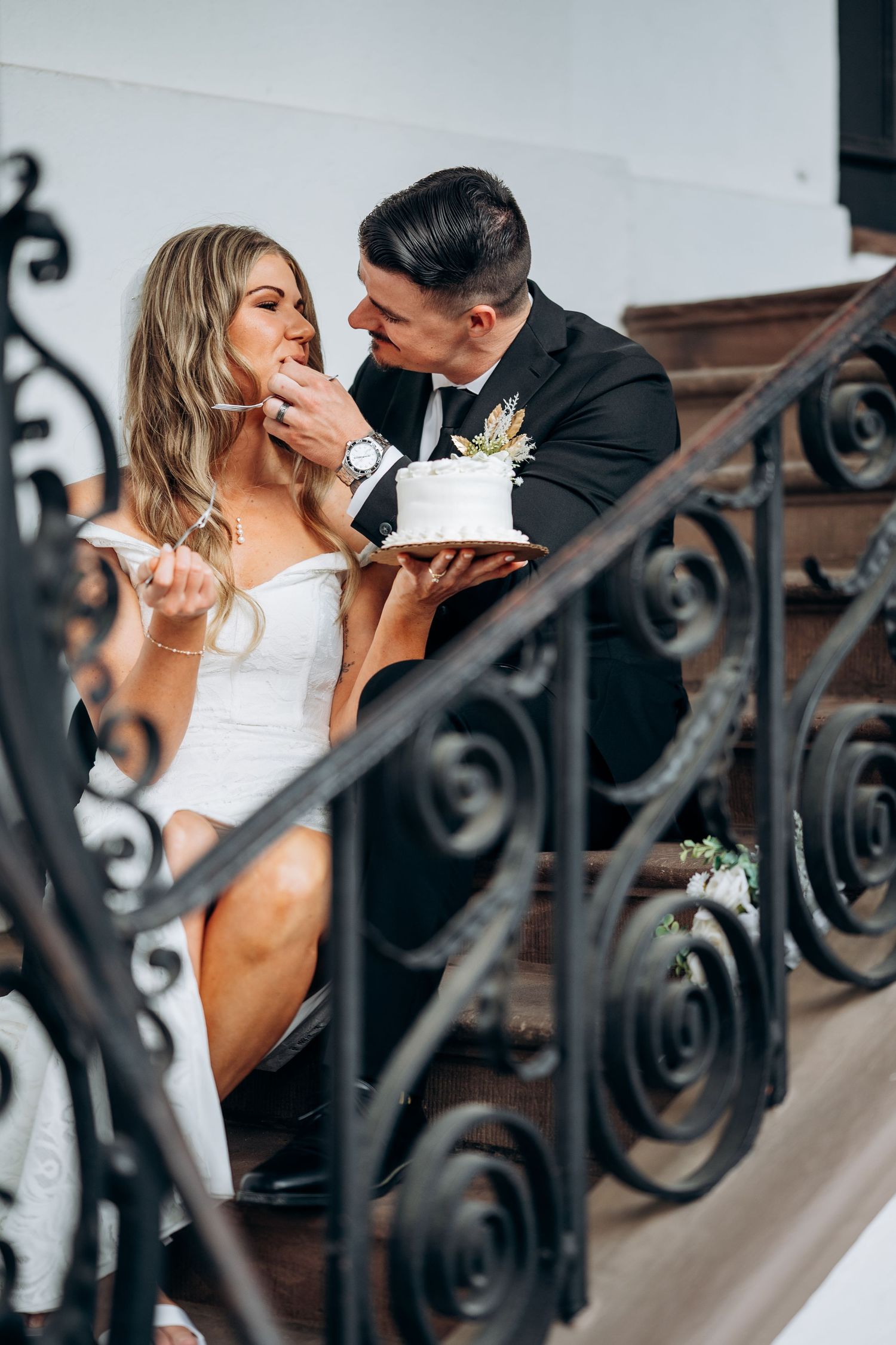 Bride and groom sharing wedding cake on ornate staircase, groom feeding bride in white dress and black suit.