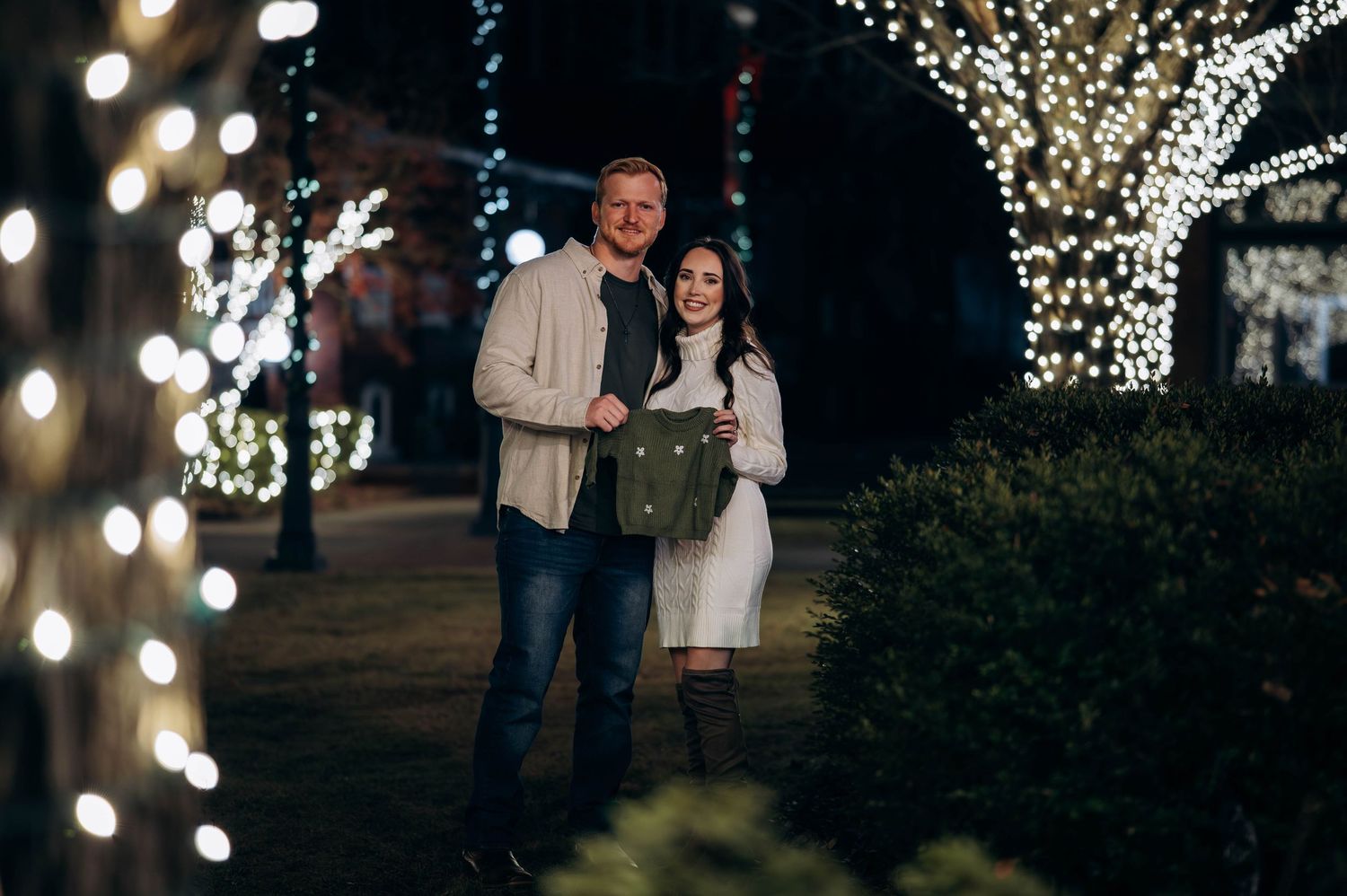 Couple smiling together outdoors at night surrounded by festive white string lights on trees.