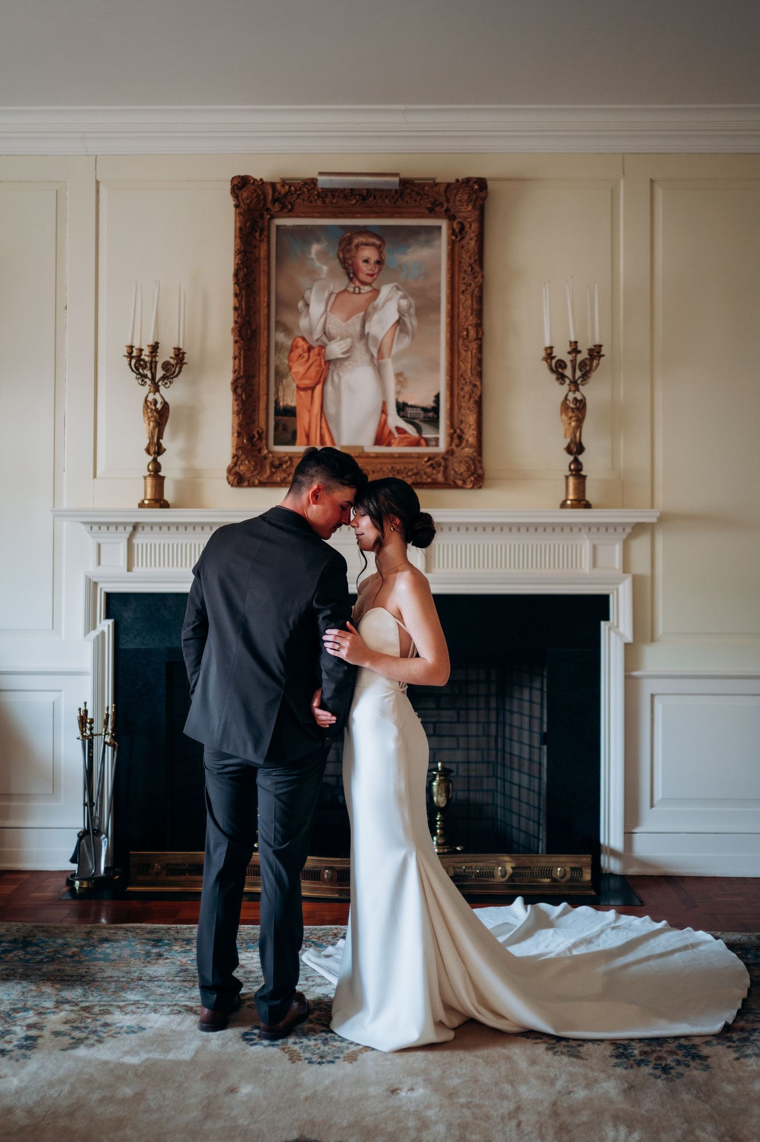 Bride and groom share an intimate moment by a fireplace beneath an ornate portrait in an elegant manor.