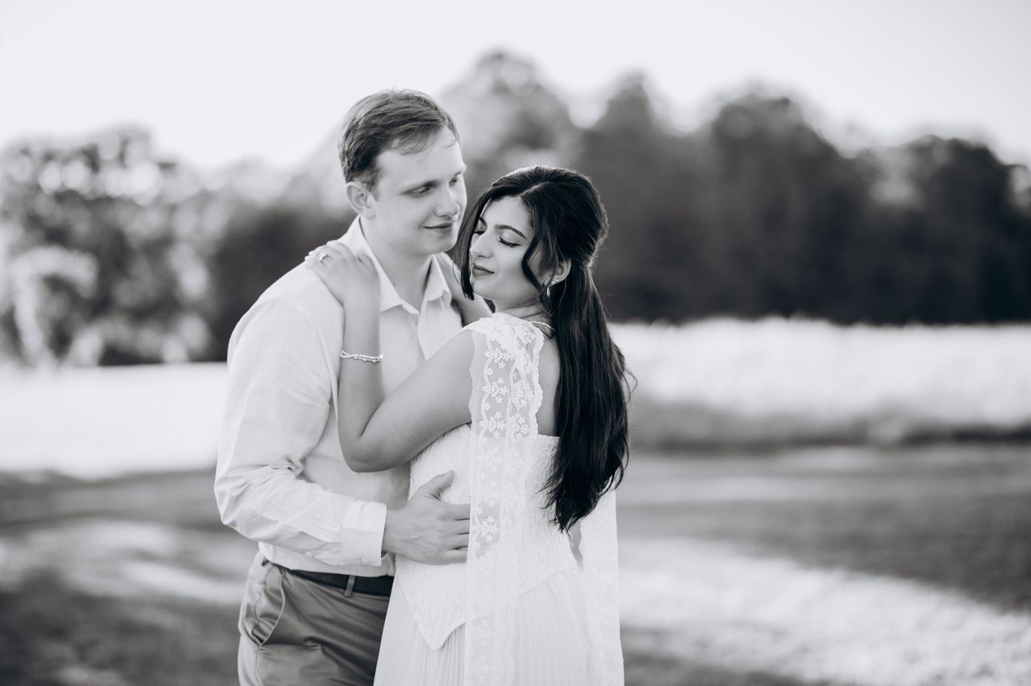 Black and white photo of a couple embracing outdoors, woman in white dress, man in white shirt, romantic pose.