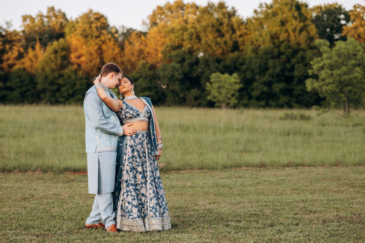 Couple embracing in a green field at golden hour, woman in blue floral lehenga, man in light blue suit.