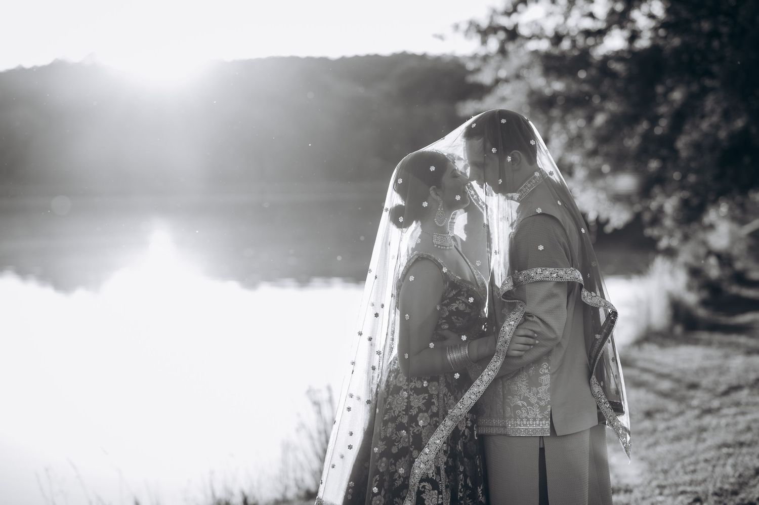 Black and white photo of a couple sharing a romantic moment under a bridal veil by a serene lake.