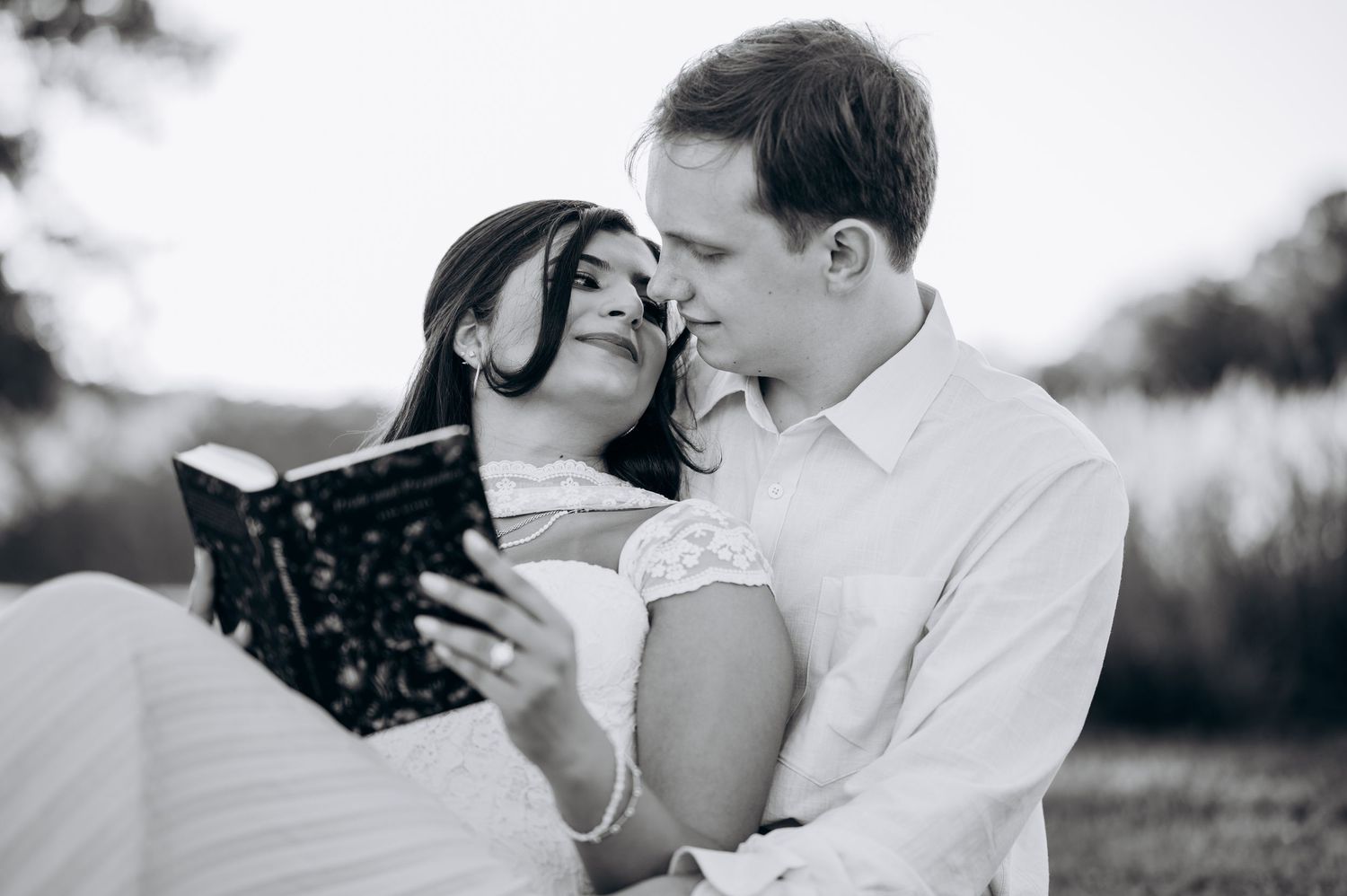 Black and white photo of a romantic couple in wedding attire sharing a tender moment while reading a book outdoors.