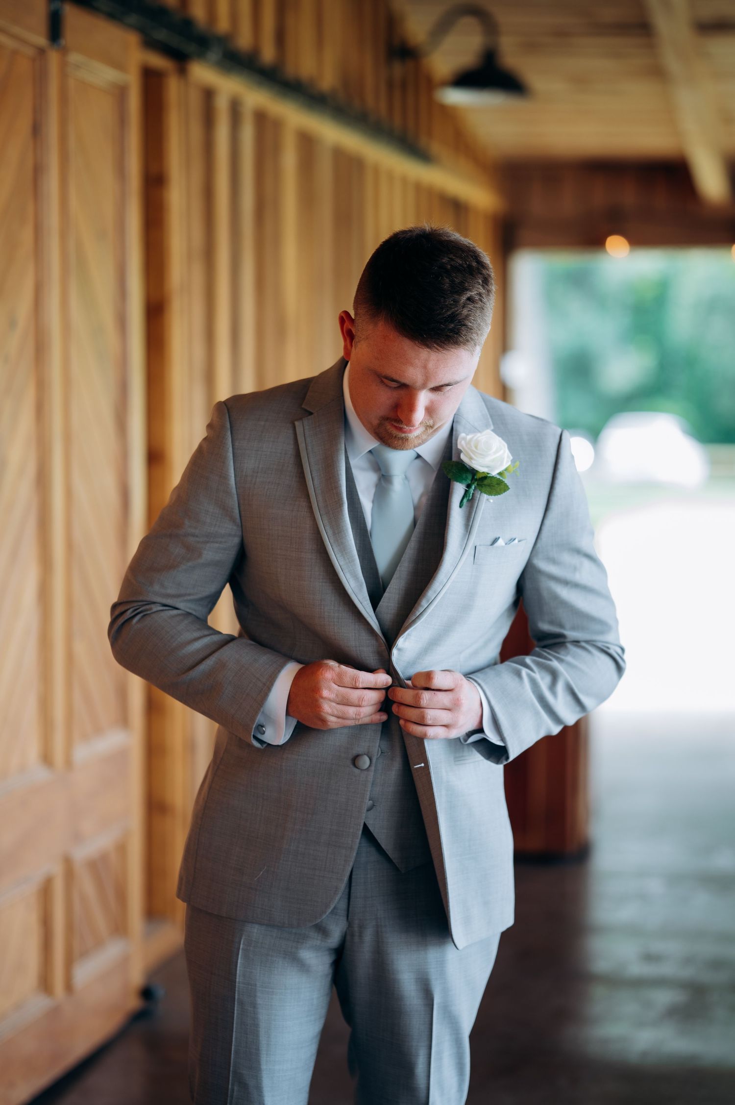 Dapper groom in gray three-piece suit with white boutonniere buttoning jacket in rustic barn venue.