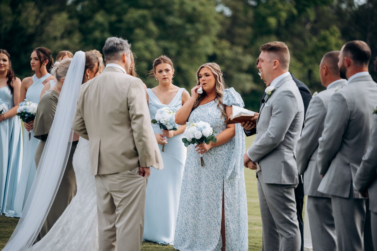 Outdoor wedding ceremony with bridesmaids in light blue dresses, groomsmen in gray suits, and officiant reading vows on green lawn.