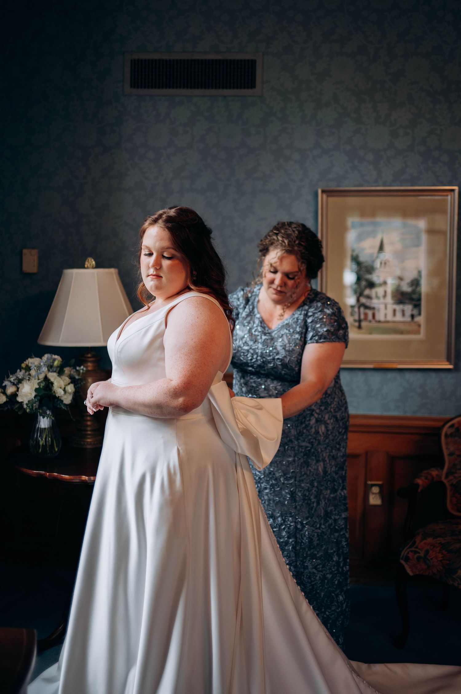 A bride in a white gown gets help with final dress adjustments in a dimly lit room.