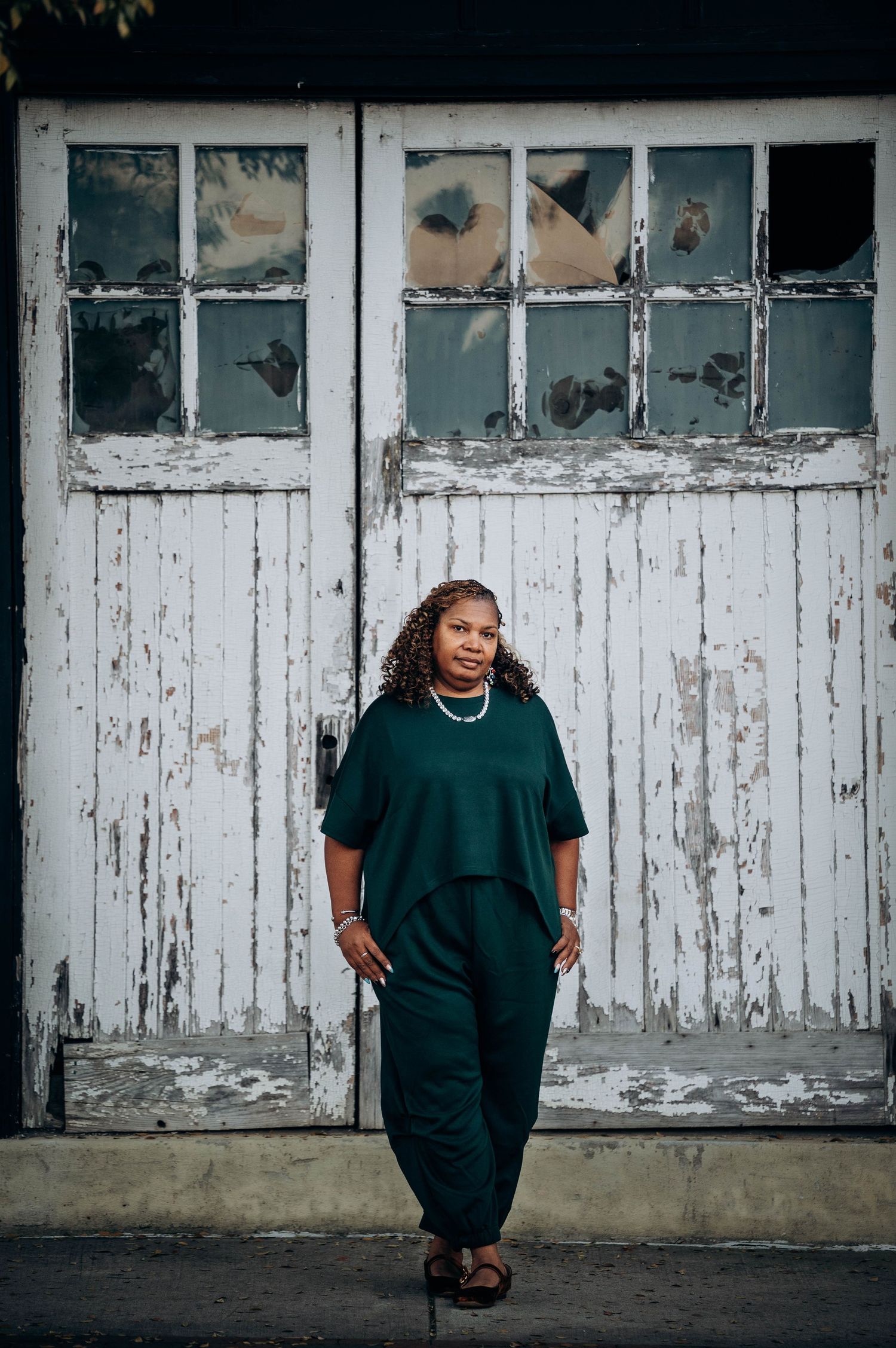 A person in green clothing poses against a weathered white door with broken windows.