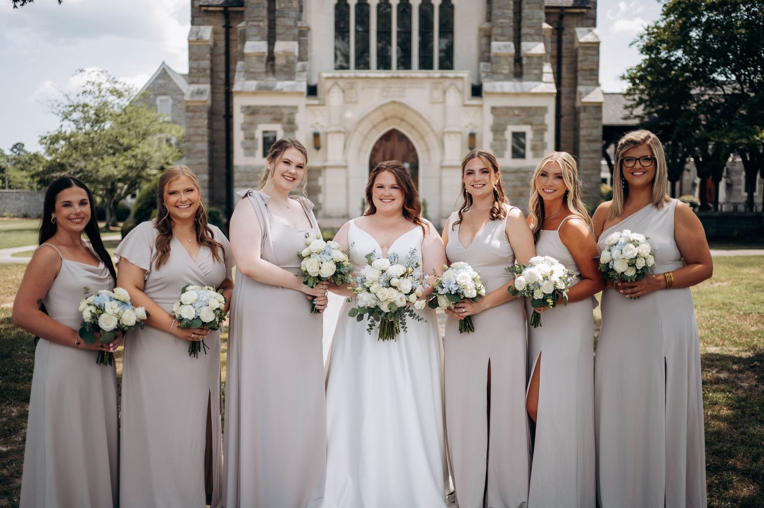 Wedding party in light gray dresses and white gowns standing outside a stone church.