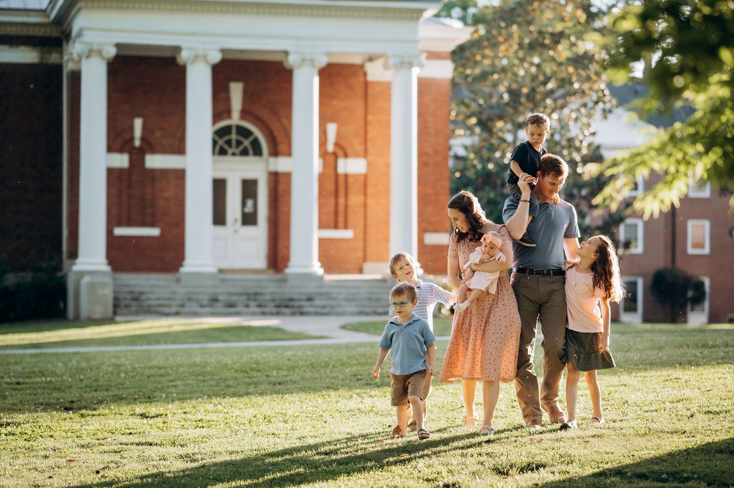 Happy family with young children walking on green lawn outside a classic red brick building on a sunny day.