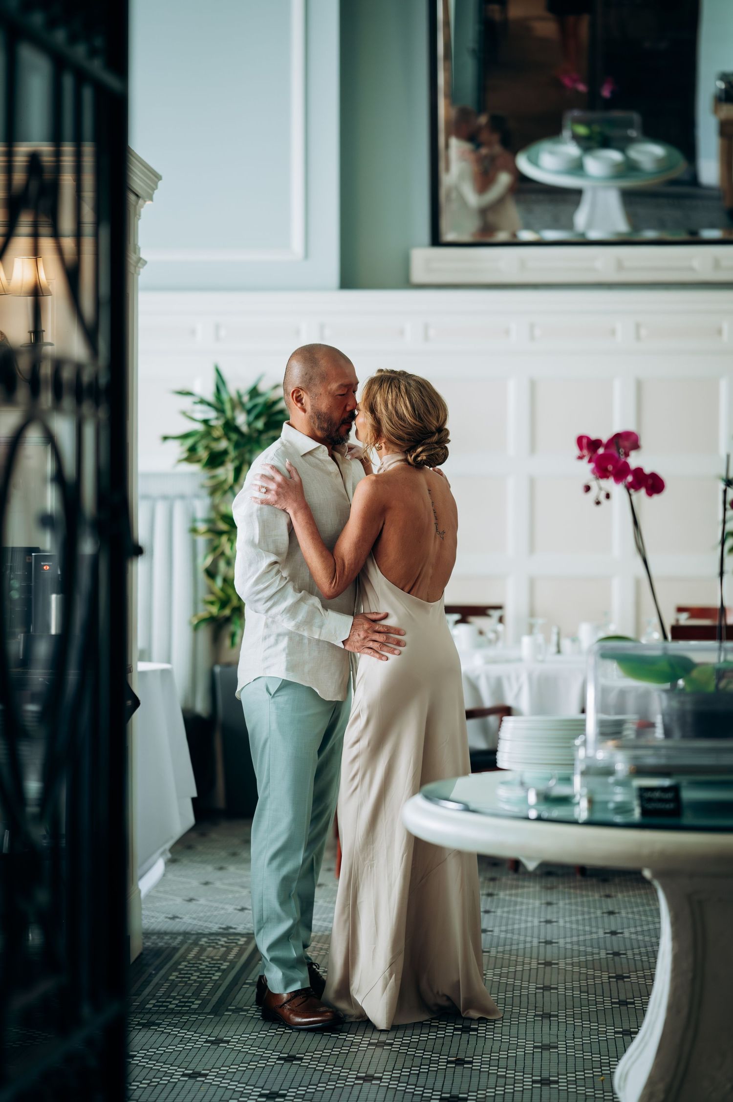 Couple sharing a romantic dance in an elegant restaurant, bride in backless gown, groom in light suit.