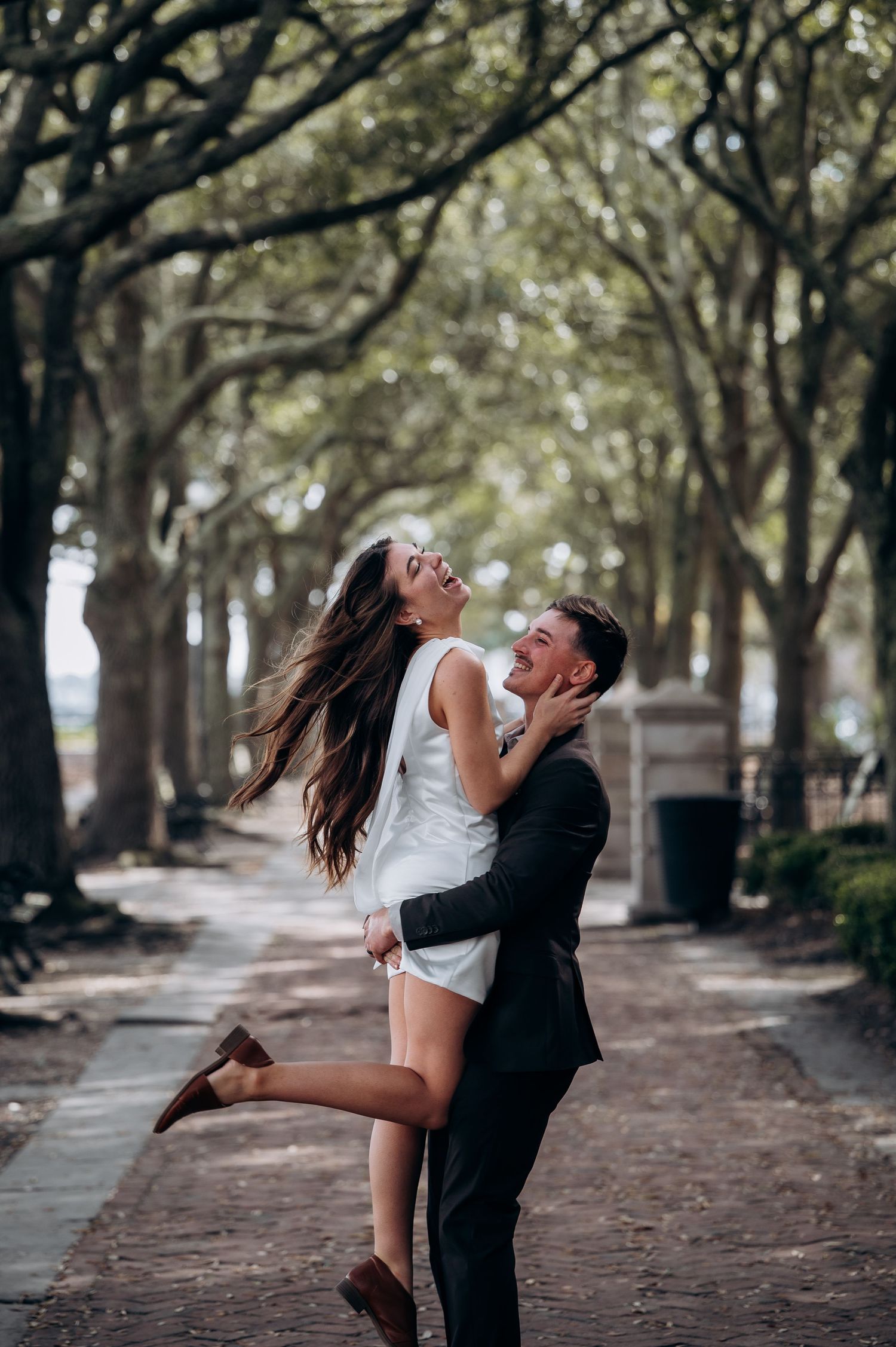 Groom joyfully lifting laughing bride along a tree-lined path on their wedding day.