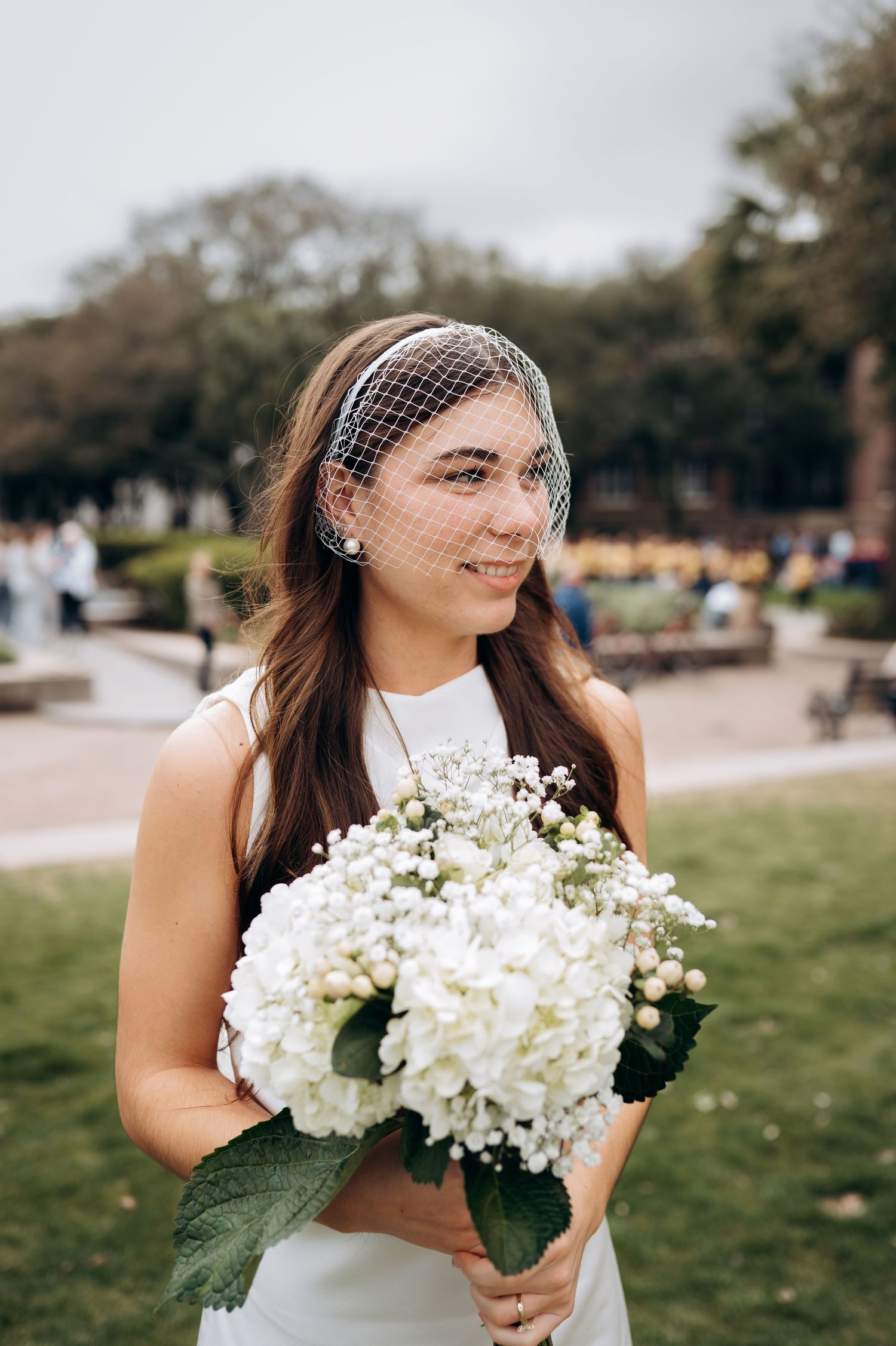 Bride in white dress holding white hydrangea bouquet wearing birdcage veil in outdoor park setting.
