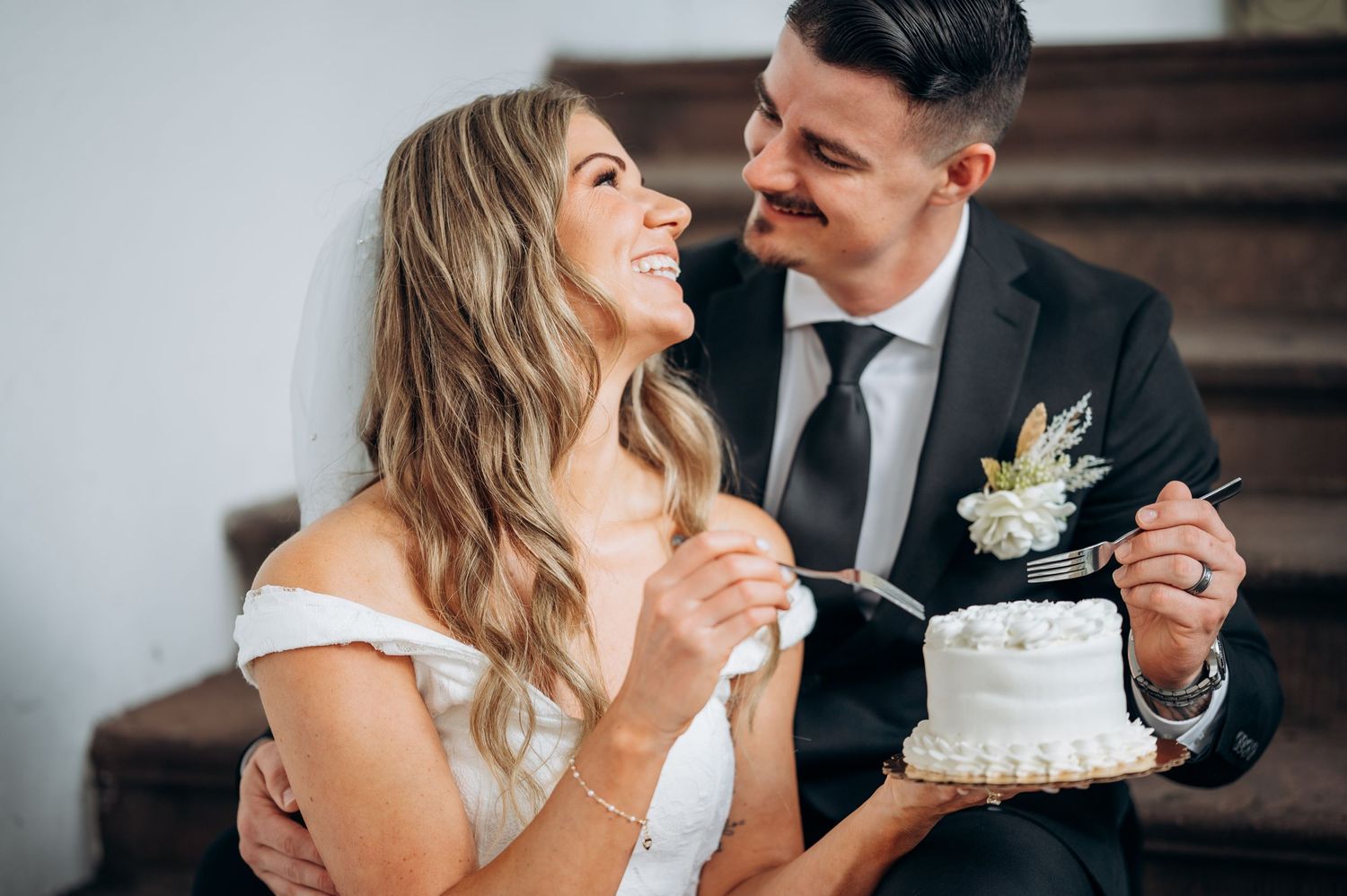 Bride and groom smiling at each other while cutting a small white wedding cake together.