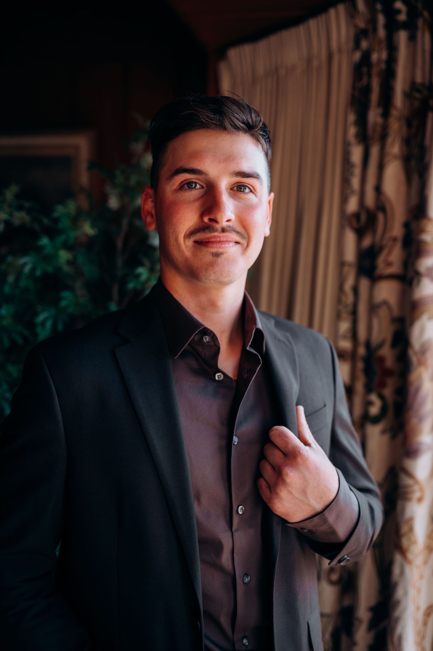 Portrait of someone in a dark suit jacket and button-up shirt standing against a curtained backdrop with foliage.