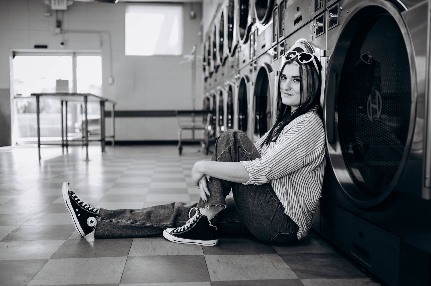 Black and white photo of someone sitting on laundromat floor against washing machines wearing casual attire.
