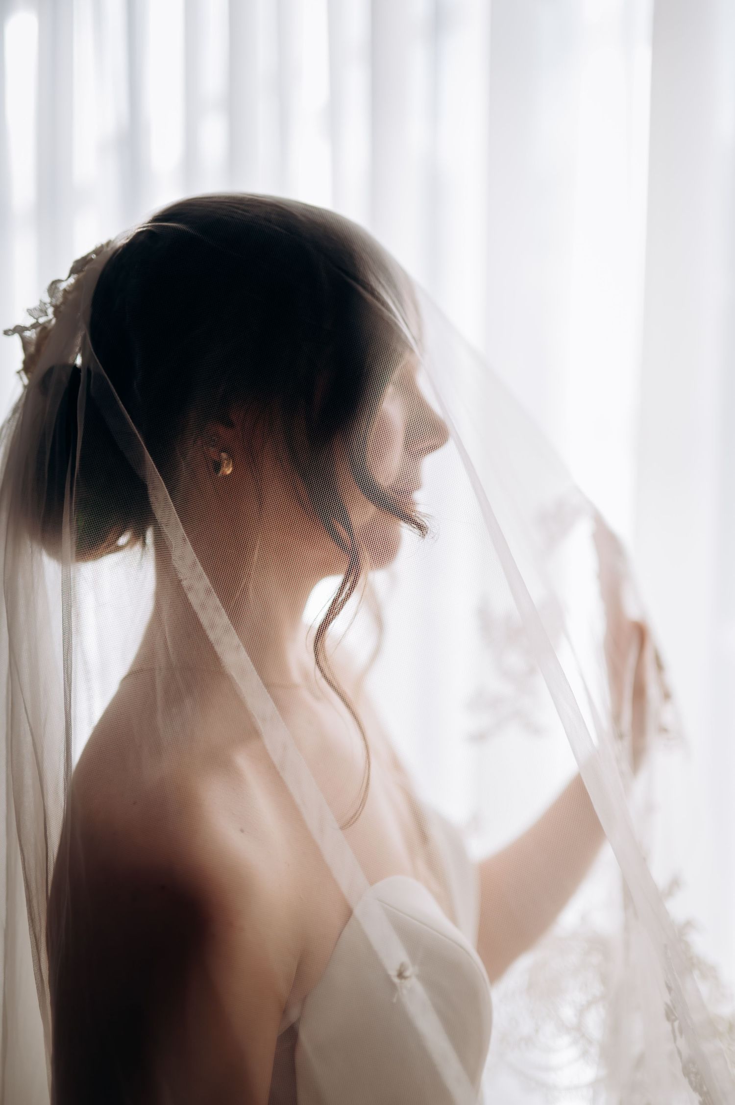 Ethereal silhouette of a bride in a veil against soft white curtains.