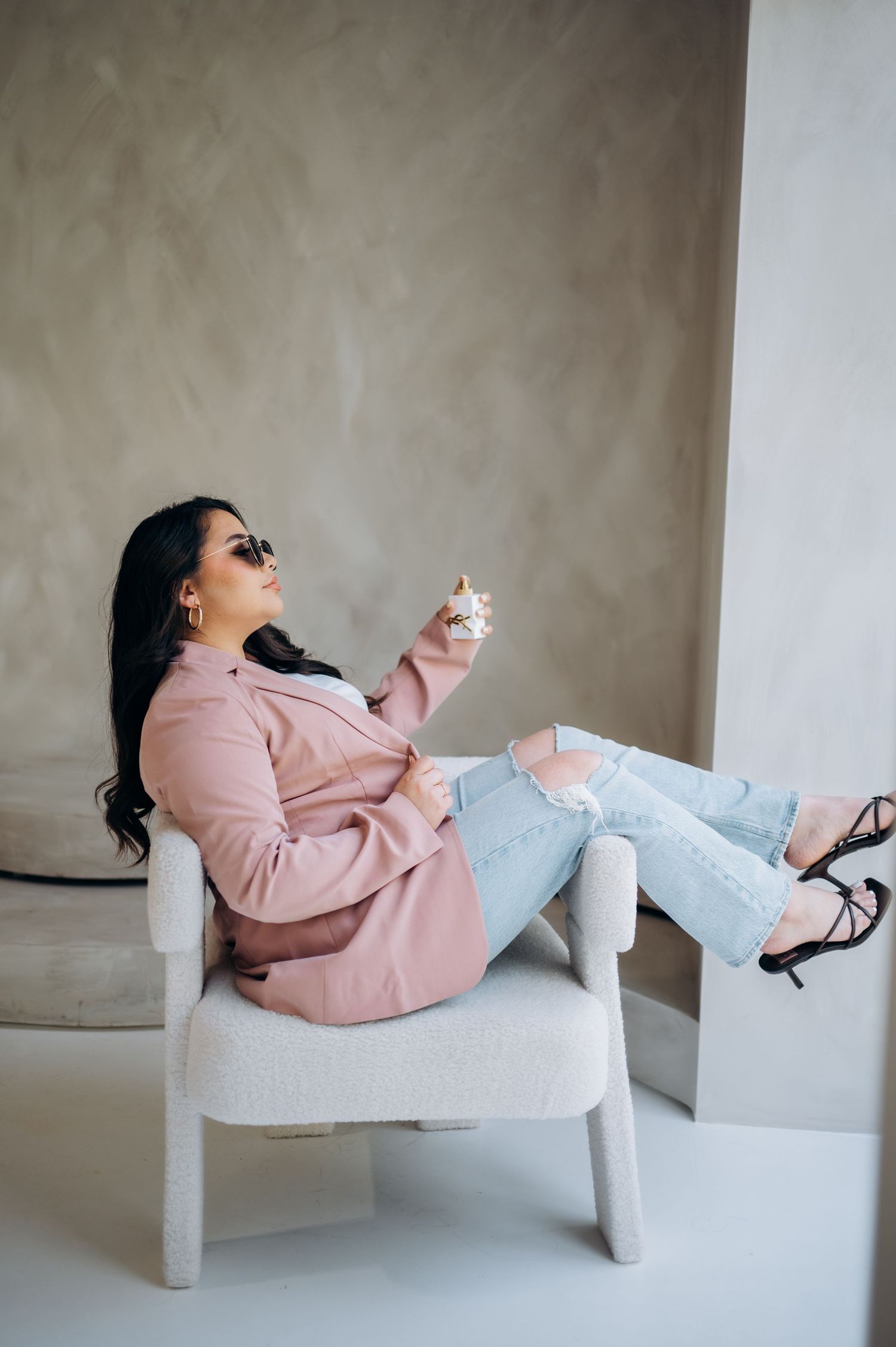 A person in a pink sweatshirt lounges and laughs while reclining on a white modern chair against a neutral wall.