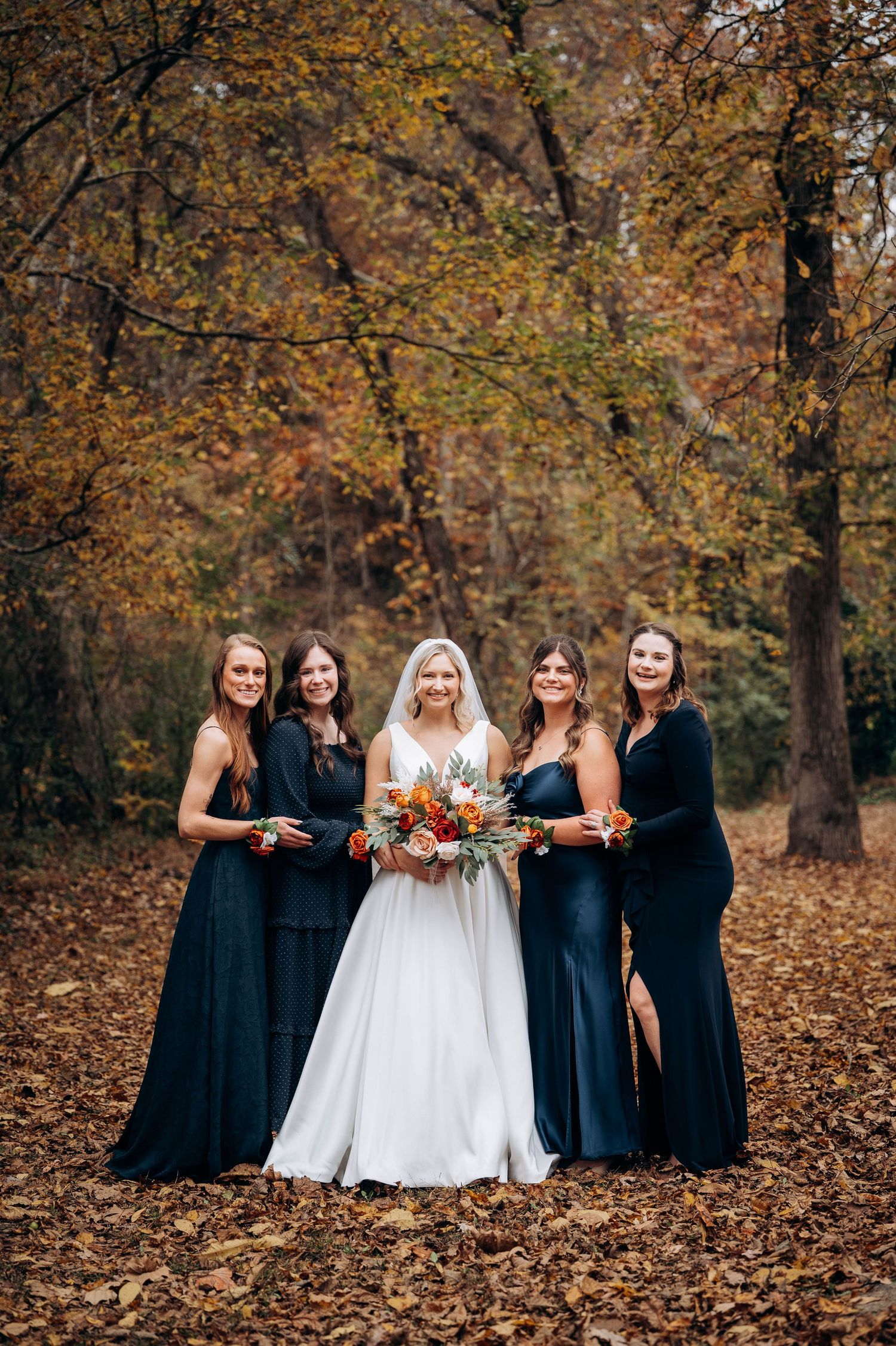 Bride in white dress poses with bridesmaids in navy dresses among autumn leaves.