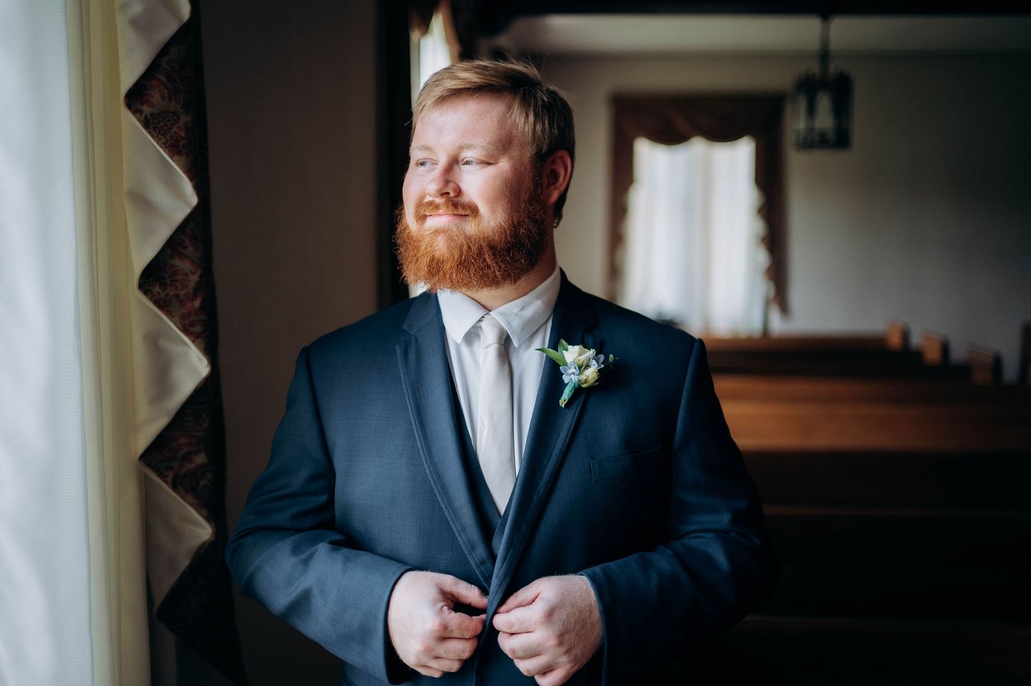 A groom in a navy suit adjusts his jacket while getting ready indoors.