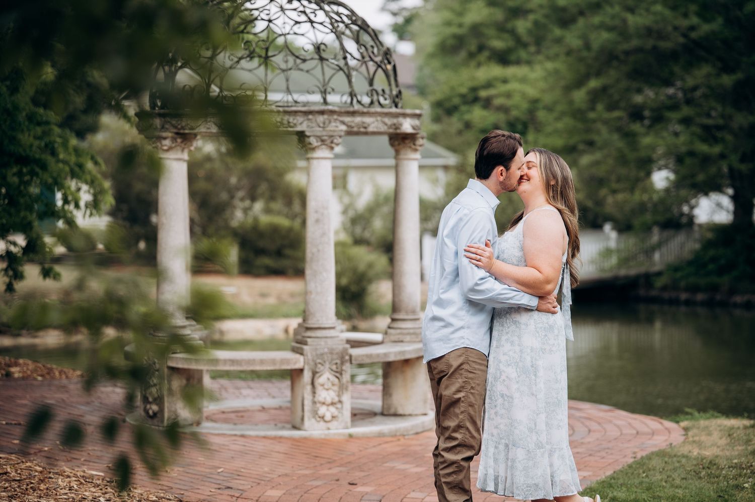 Couple kissing in front of elegant stone gazebo beside a pond in a garden setting.