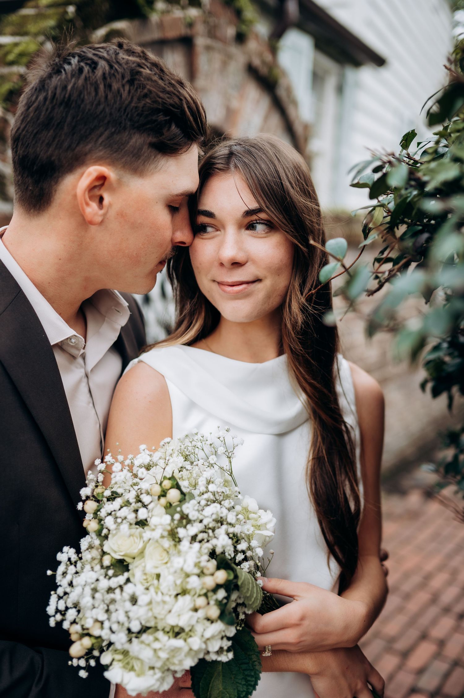 Bride smiles softly as groom nuzzles her cheek, she holds a white bouquet of hydrangeas and baby's breath.