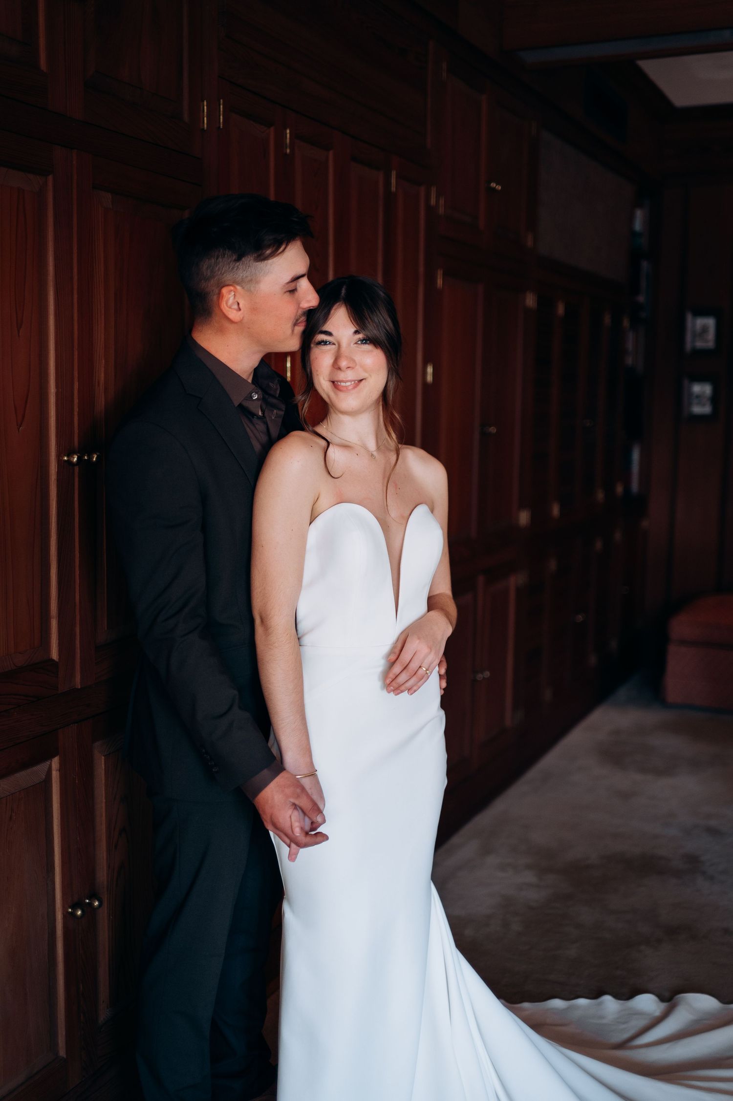Groom in dark suit embraces smiling bride in white mermaid gown against rich wood-paneled wall interior.