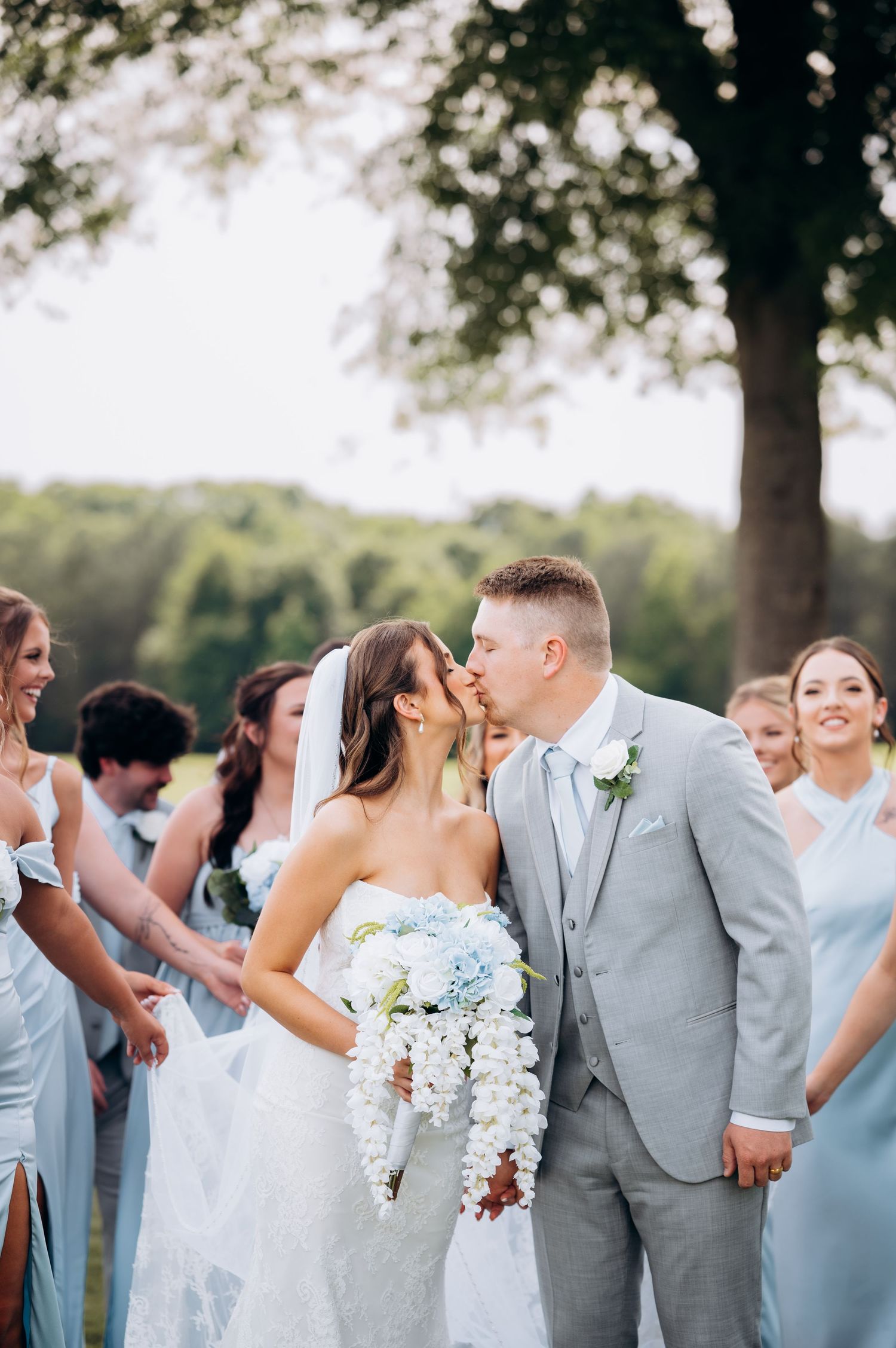 Bride and groom share a kiss surrounded by bridesmaids in blue dresses and groomsmen in gray suits outdoors.
