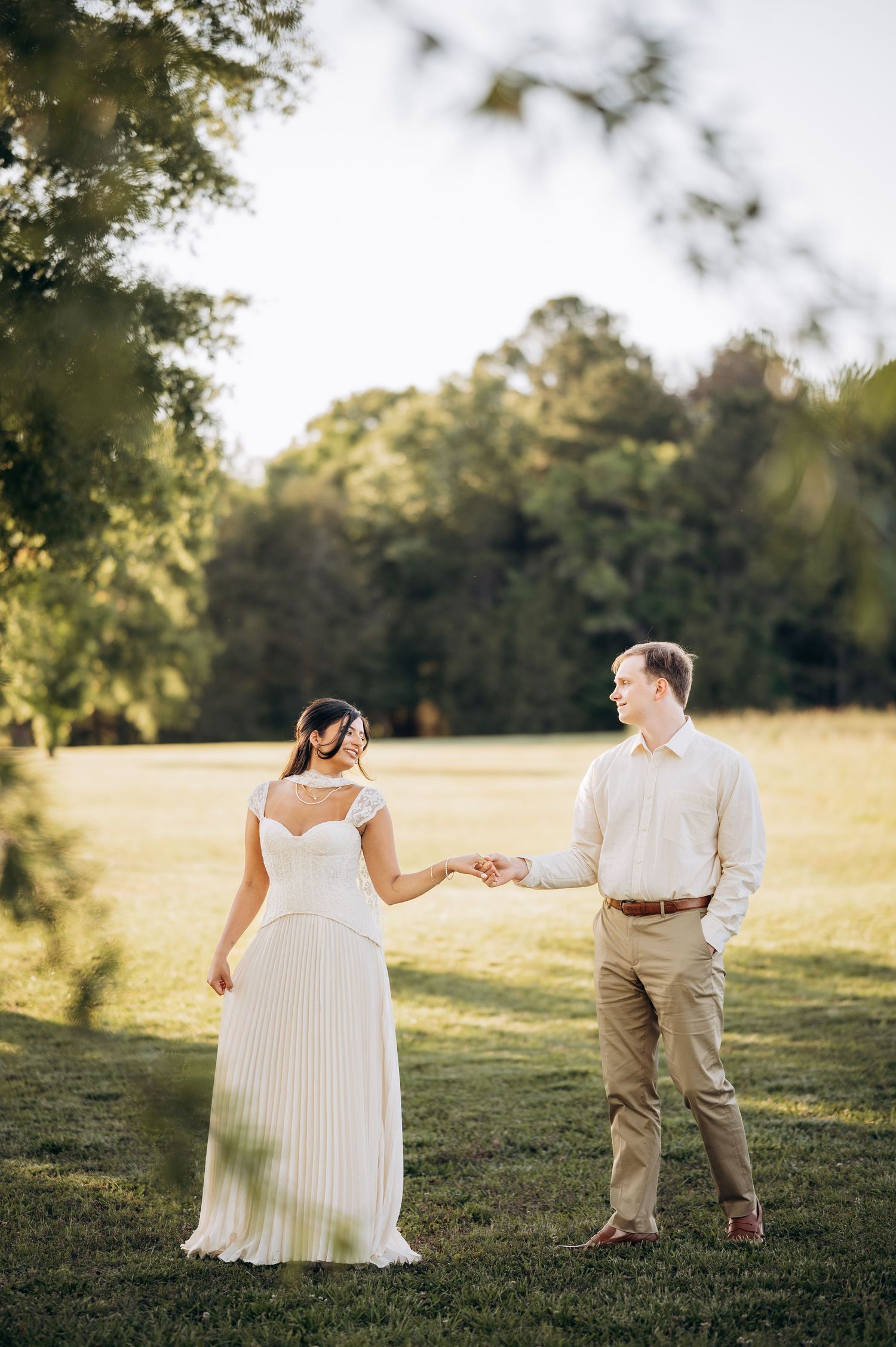 Bride in white pleated gown holds hands with groom in khaki pants on lush green lawn surrounded by trees.