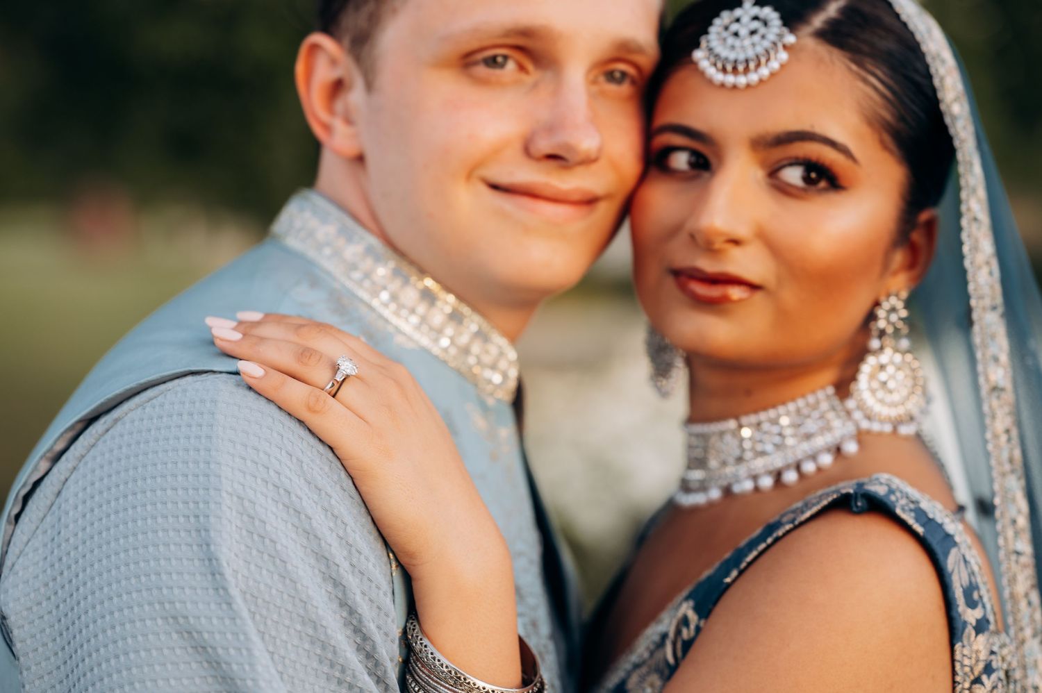 Interracial couple in wedding attire, bride wearing traditional Indian jewelry and blue lehenga, groom in light blue suit.
