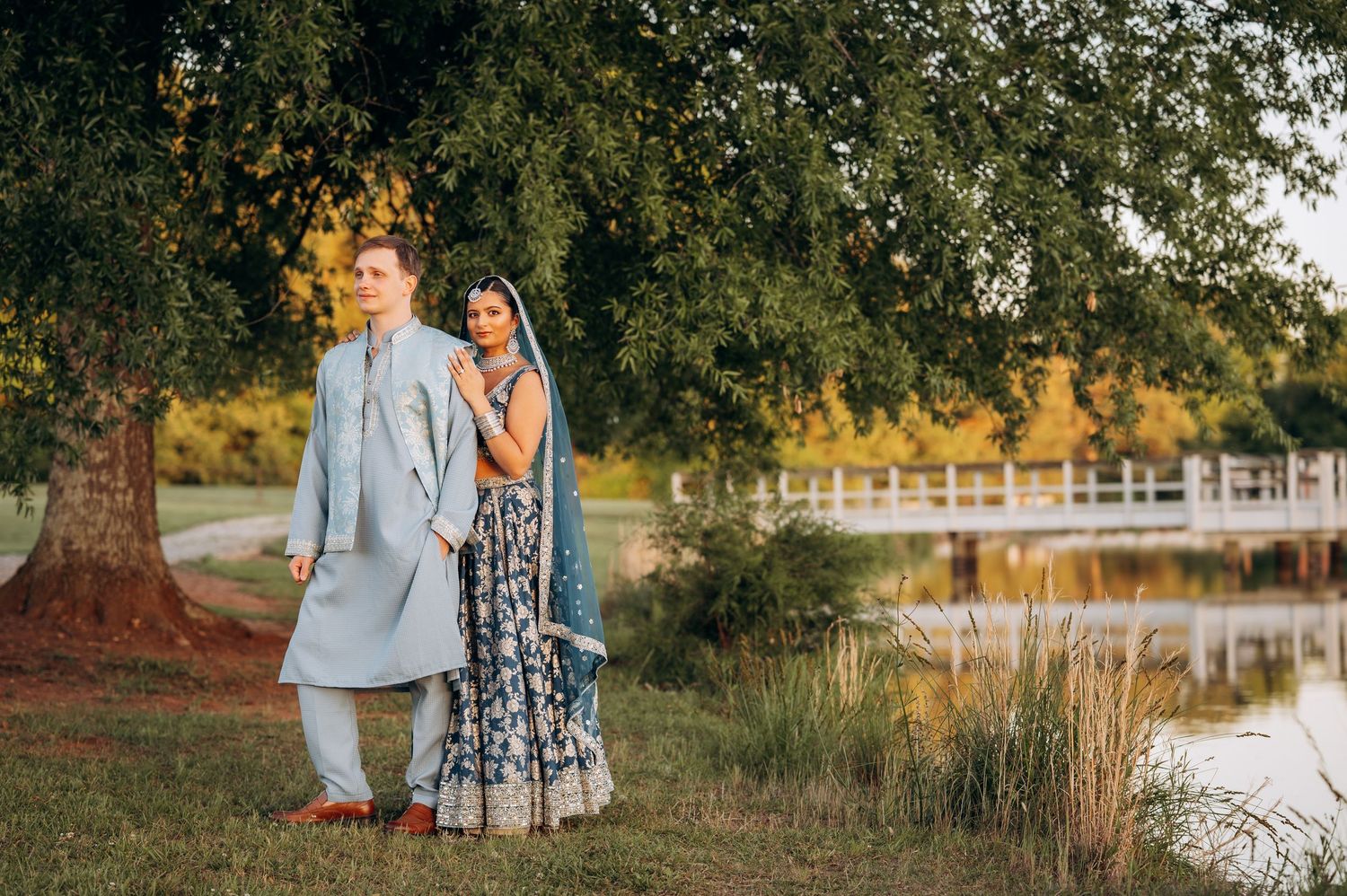 Couple in traditional Indian wedding attire standing by a serene pond under a large tree at golden hour.