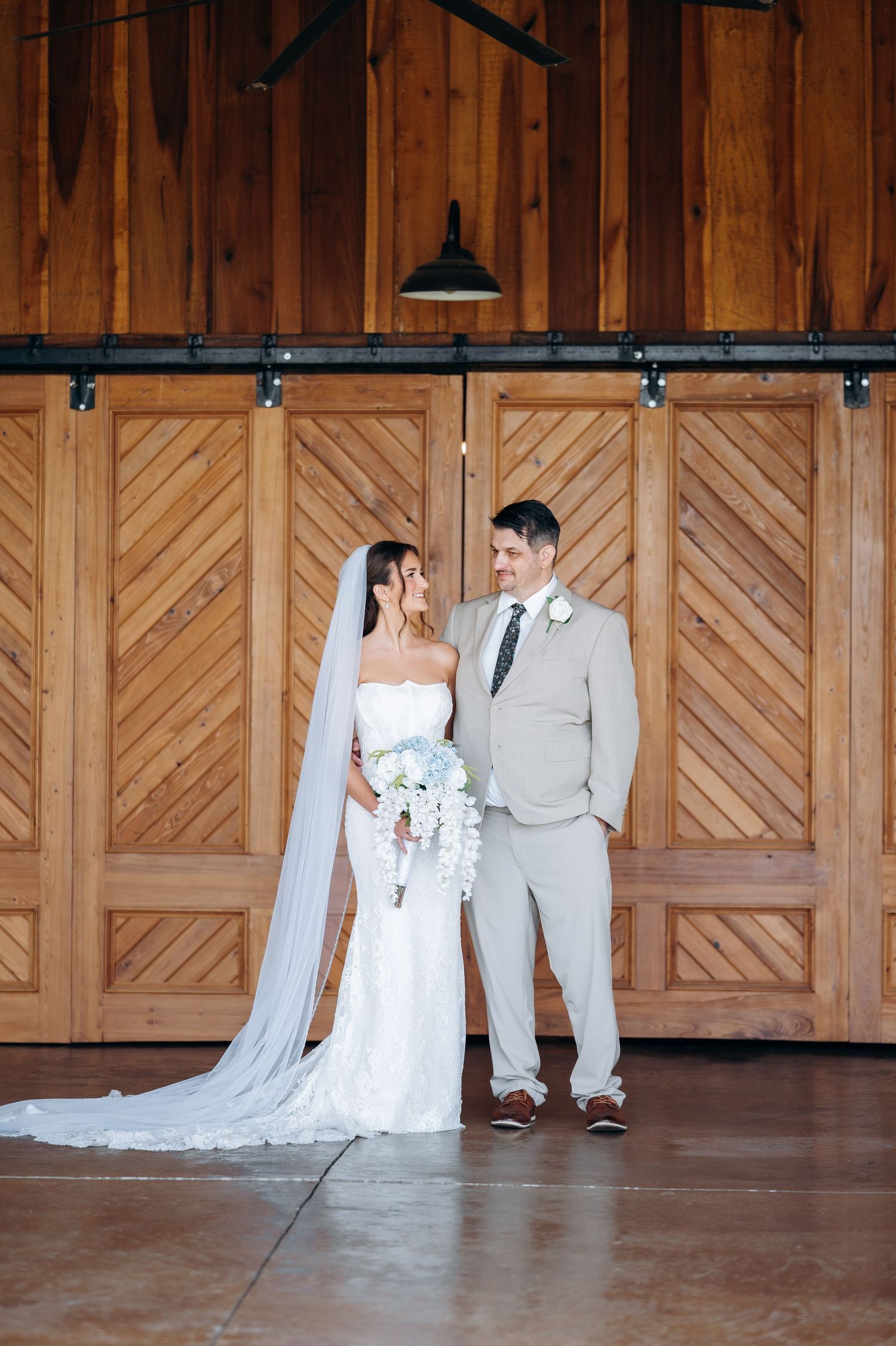 Bride in white gown with veil and groom in tan suit pose before rustic wooden barn doors at wedding.