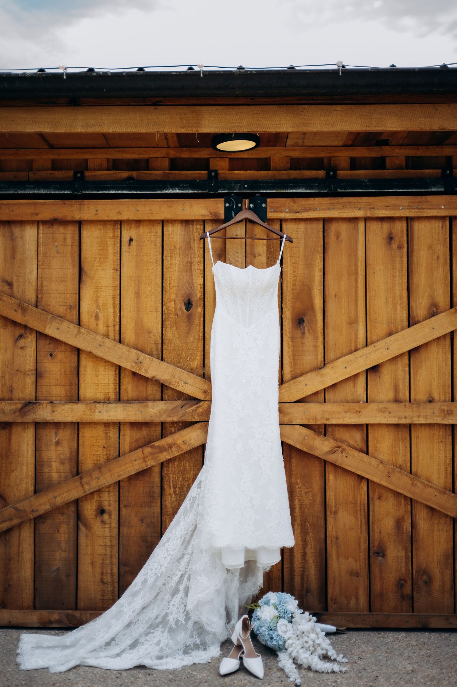 White lace wedding dress hanging on wooden barn doors with bridal bouquet and white heels below.